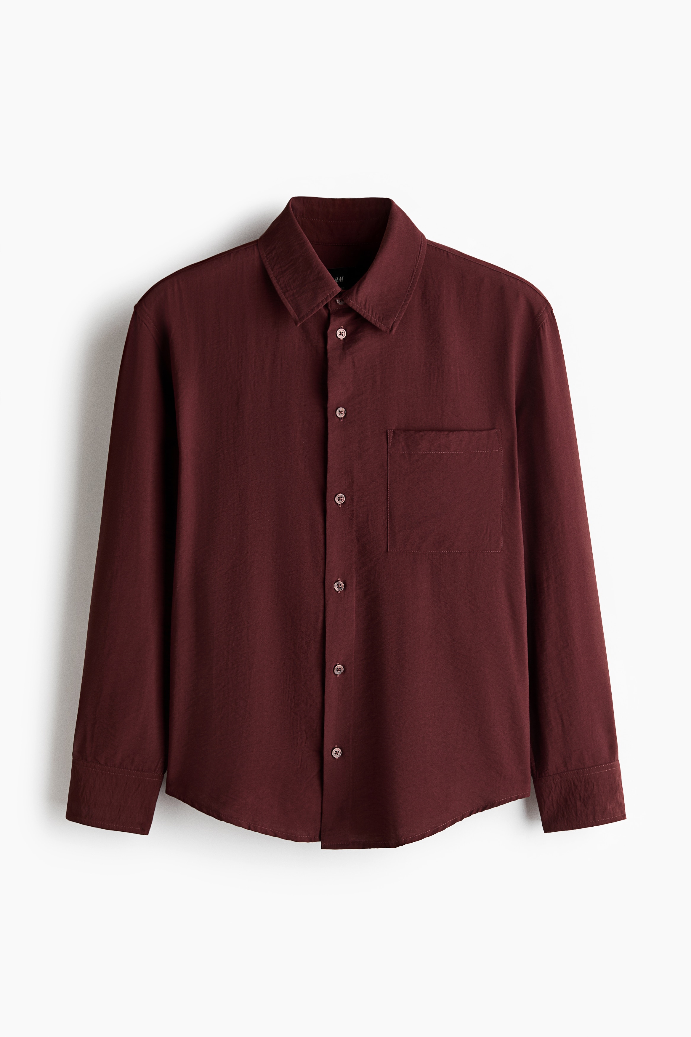 A burgundy long-sleeve shirt in a lightly crinkled fabric is worn by the model, tucked into dark trousers with a black belt. The relaxed-fit shirt features a collar, a partially unbuttoned front placket, and a single patch chest pocket.