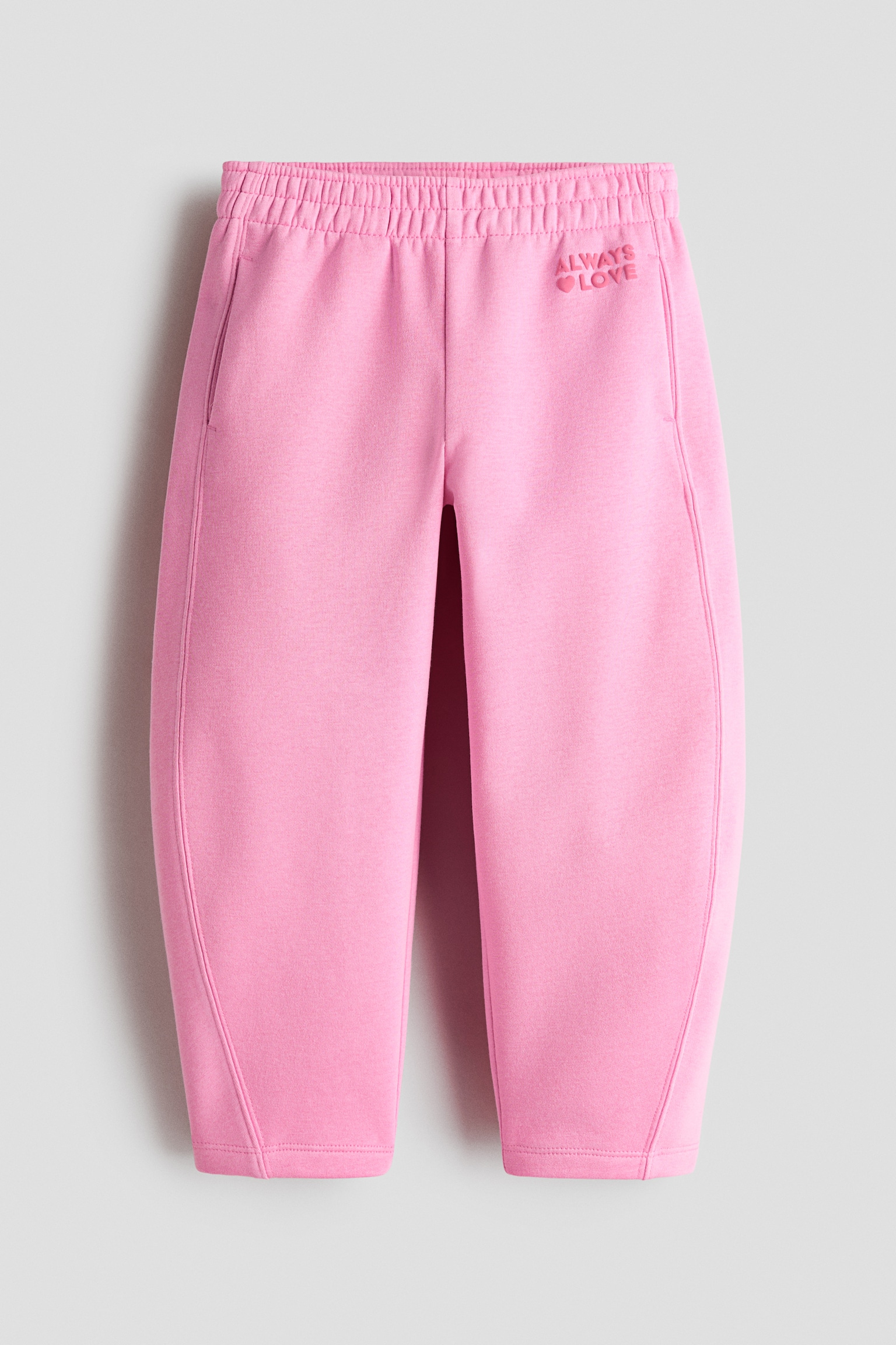 Ein Paar rosa Sweatpants ist von vorne zu sehen und präsentiert einen elastischen Bund, Seitentaschen und eine leicht geschwungene Beinsilhouette. Am oberen linken Oberschenkel ist tonale Stickerei mit dem Schriftzug âALWAYSâ über einem kleinen Herzsymbol und dem Wort âLOVEâ zu sehen.