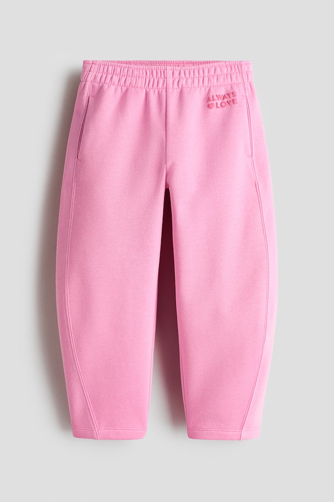 A pair of pink sweatpants is shown from the front, featuring an elasticised waistband, side pockets, and a slightly curved leg silhouette. The upper left thigh features tonal embroidered text reading "ALWAYS" above a small heart icon and the word "LOVE." - 1