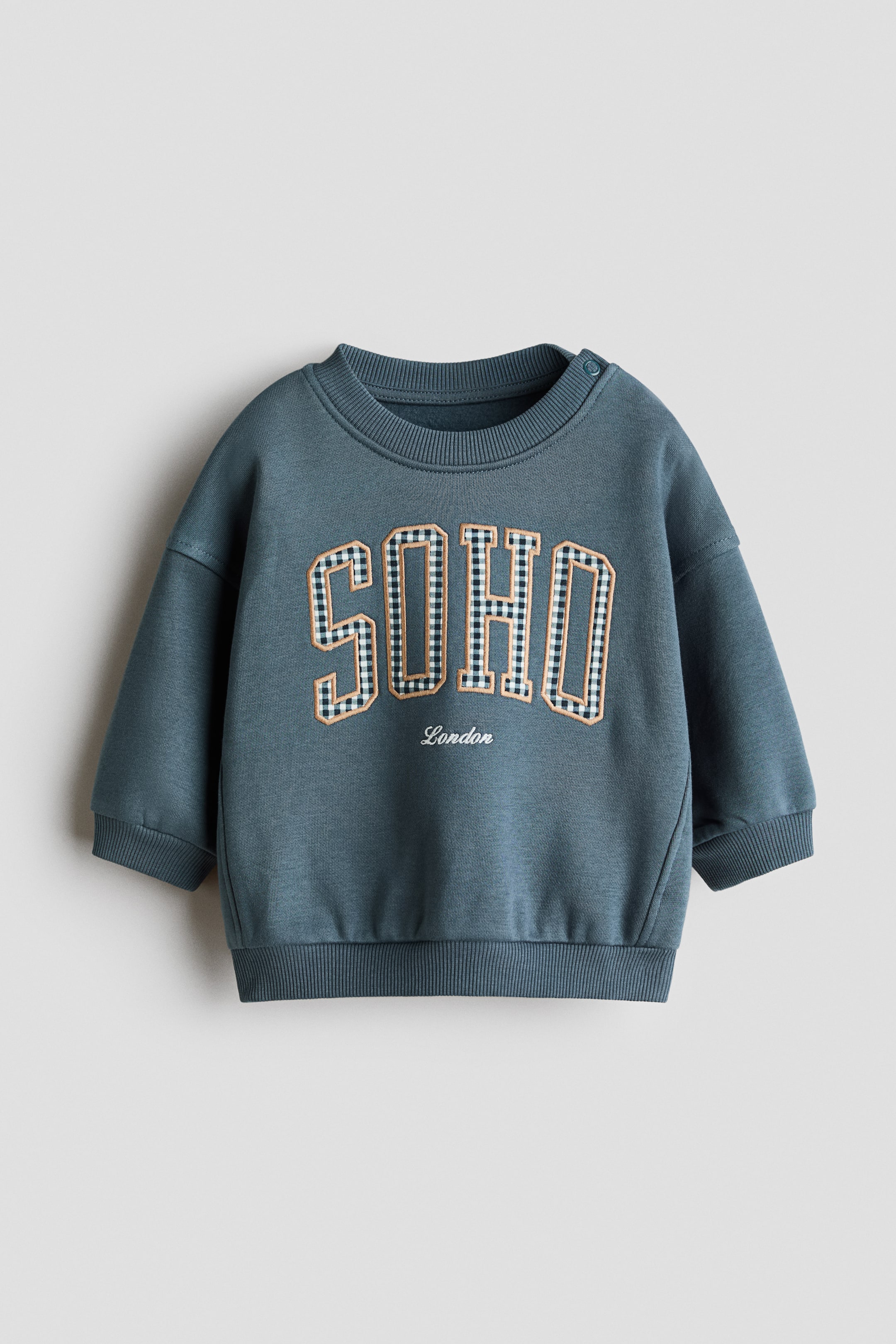 Visualizza l'immagine ingrandita: Una felpa con dettagli a motivo si presenta frontalmente in un colore teal, con "SOHO" in lettere a quadretti bianche e nere contornate in beige, con "London" ricamato sotto. È progettata con un girocollo a coste, due bottoni a pressione sulla spalla sinistra, polsini a coste e un orlo a coste.
