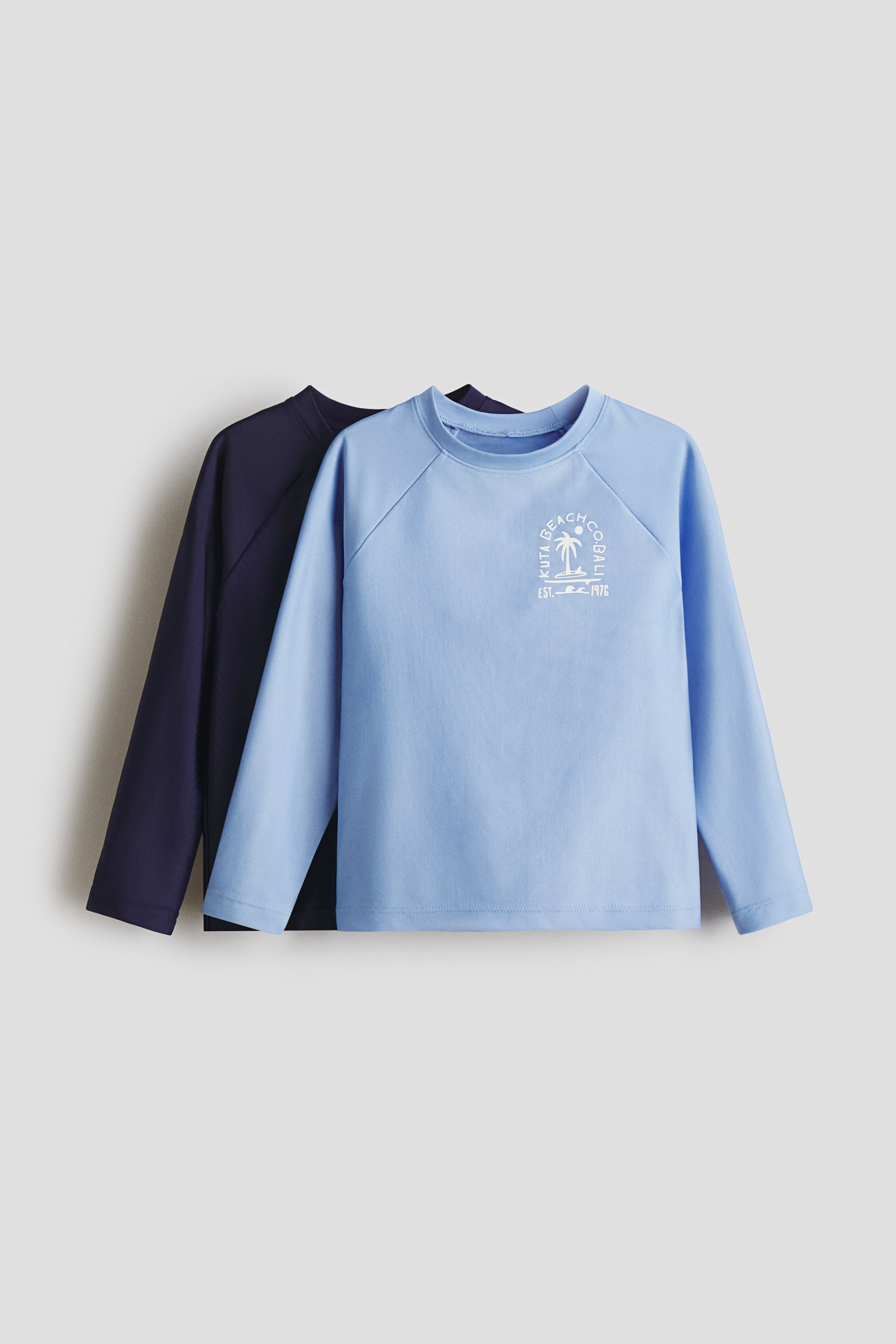 Grösseres Bild ansehen: Zwei langärmelige Oberteile sind von vorne zu sehen, wobei ein hellblaues Oberteil über einem dunkelblauen Oberteil liegt. Das hellblaue Oberteil zeigt einen weißen Grafikdruck auf der Brust, der eine Palme, Sonne, ein Surfbrett und den Text "KUTA BEACH CO. BALI EST. 1976" darstellt und ist mit Raglanärmeln und Rundhalsausschnitt designt, während das dunkelblaue Oberteil ebenfalls einen Rundhalsausschnitt aufweist.