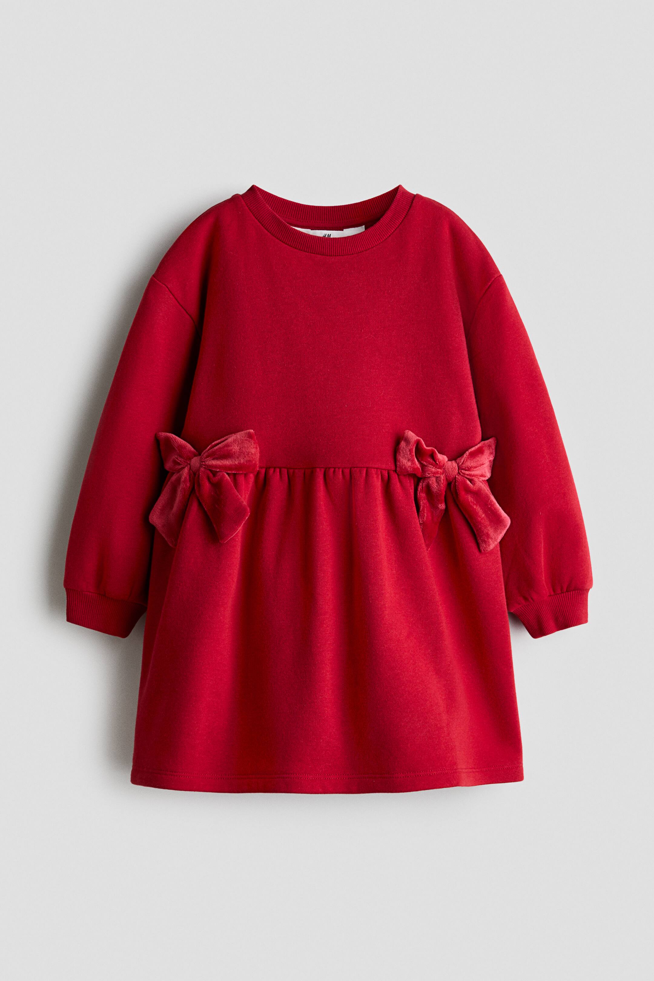 Se større billede: En rød sweatshirtkjole med lange ærmer vises forfra og har en ribbet rund hals og ribbede manchetter. Kjolen har en rynket talje, pyntet med to matchende røde fløjlsbånd i siderne, som går over i et let udsvajet skørt.