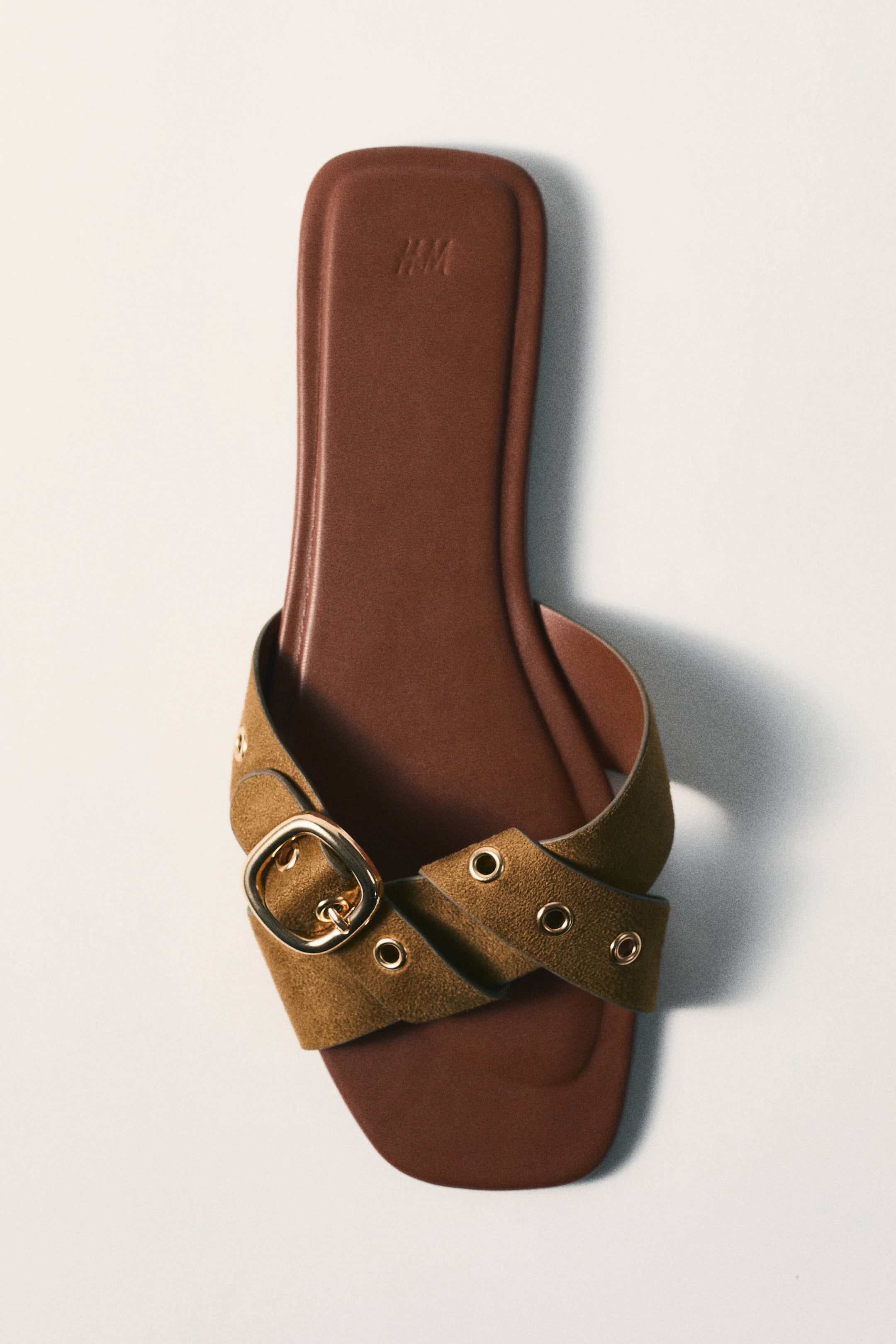 Vis større bilde: En enkel slip-in sandal ligger flatt, vinklet litt mot øvre høyre hjørne. Den har en brun fotseng med et preget H&M-logo og en bred olivengrønn stropp i semsket skinn-lignende materiale. Stroppen krysser over vristen, pyntet med et gullfarget spenne og flere gullfargede maljer.