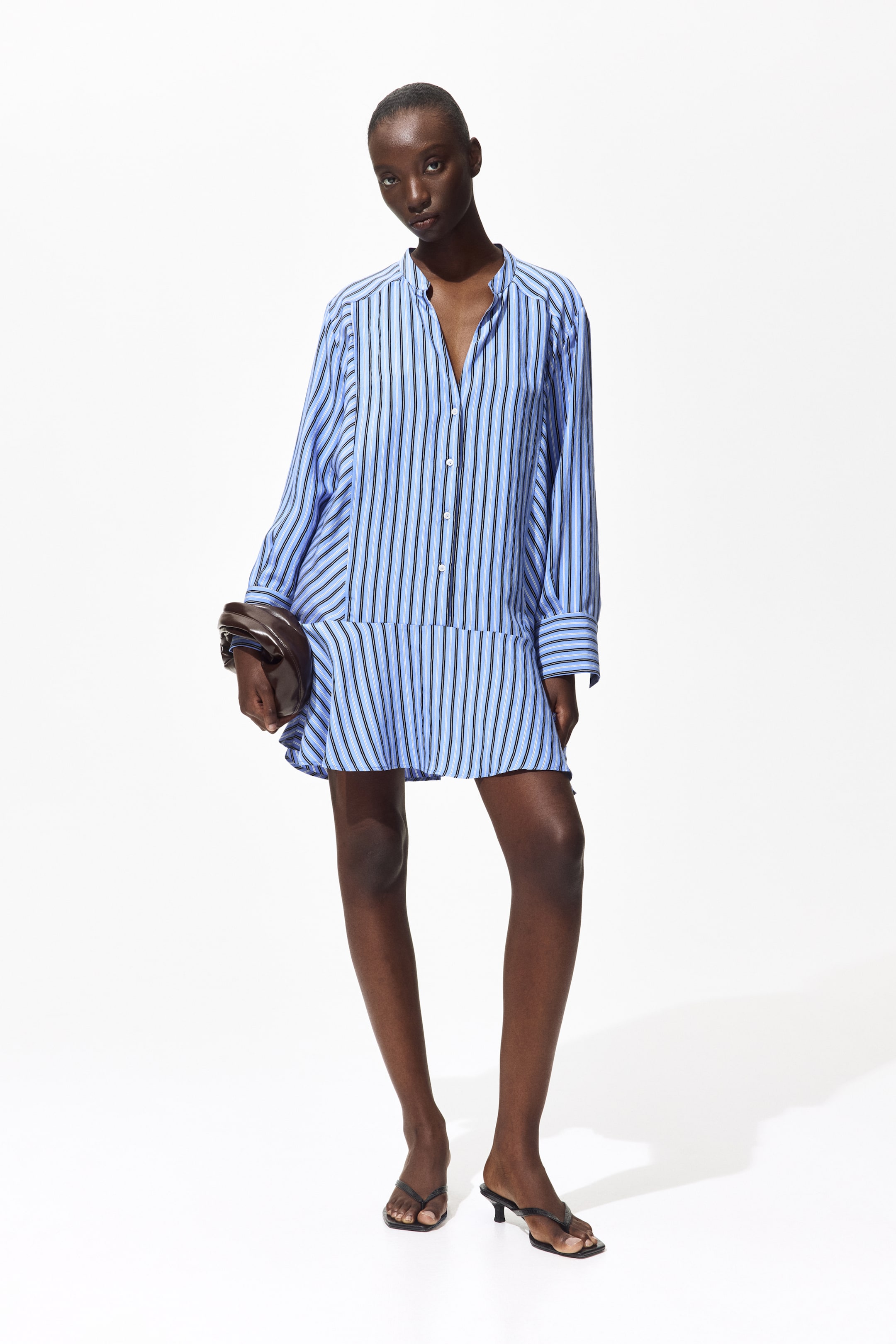 Ver imagen más grande: La modelo lleva una camisa larga tipo "popover" en un tejido de mezcla de viscosa con rayas azules y negras, que presenta un cuello tipo banda con una abertura en forma de V, una tapeta de botones alargada, costuras de hombro hacia adelante, mangas largas con puños abotonados, paneles laterales y un amplio volante en el dobladillo. La modelo sostiene un bolso de mano marrón junto a la camisa.