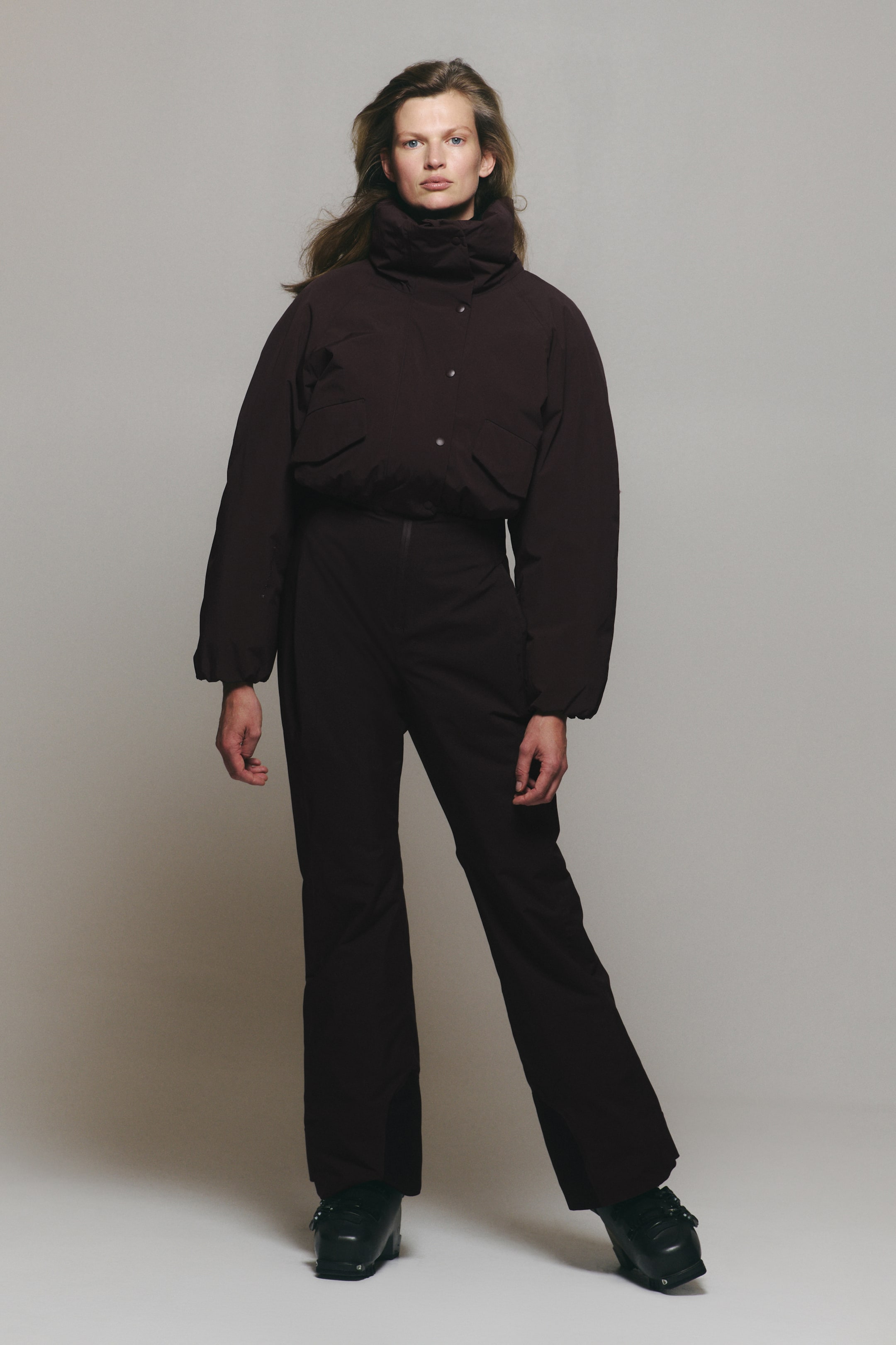 Agrandir l'image: Un ensemble de ski marron foncé, composé d’une doudoune courte avec un col montant à effet entonnoir et d’un pantalon taille haute assorti, est porté par le mannequin. La veste présente une fermeture pressionnée sur le devant et des poches à rabat, et est associée à des bottes de ski noires.