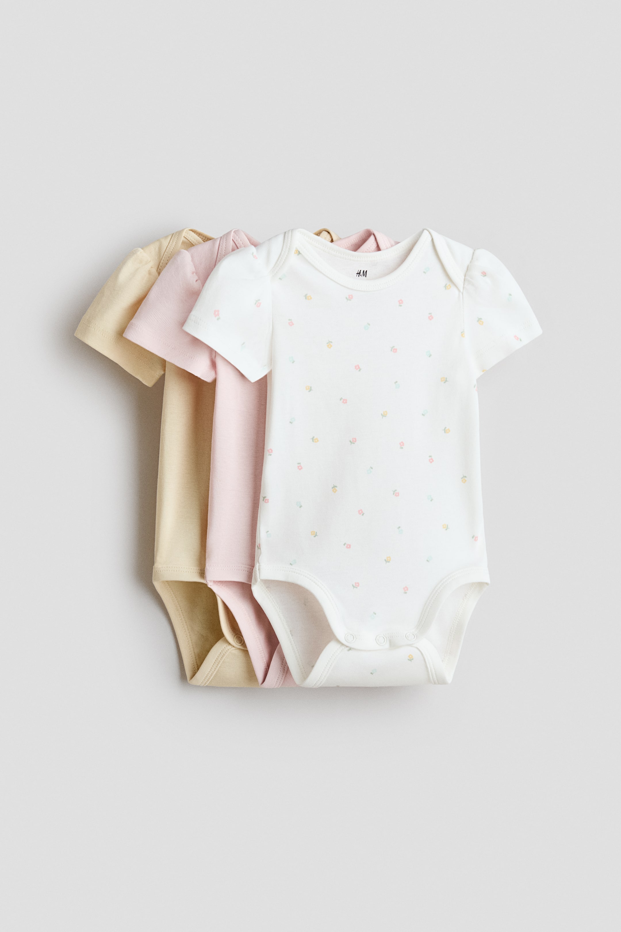 View larger image: Three baby bodysuits are layered, with the front white bodysuit facing front. It features short sleeves, a round neckline, a scattered light floral print, and snap fasteners at the shoulder and crotch. A light pink bodysuit and a light yellow bodysuit are partially visible behind it.