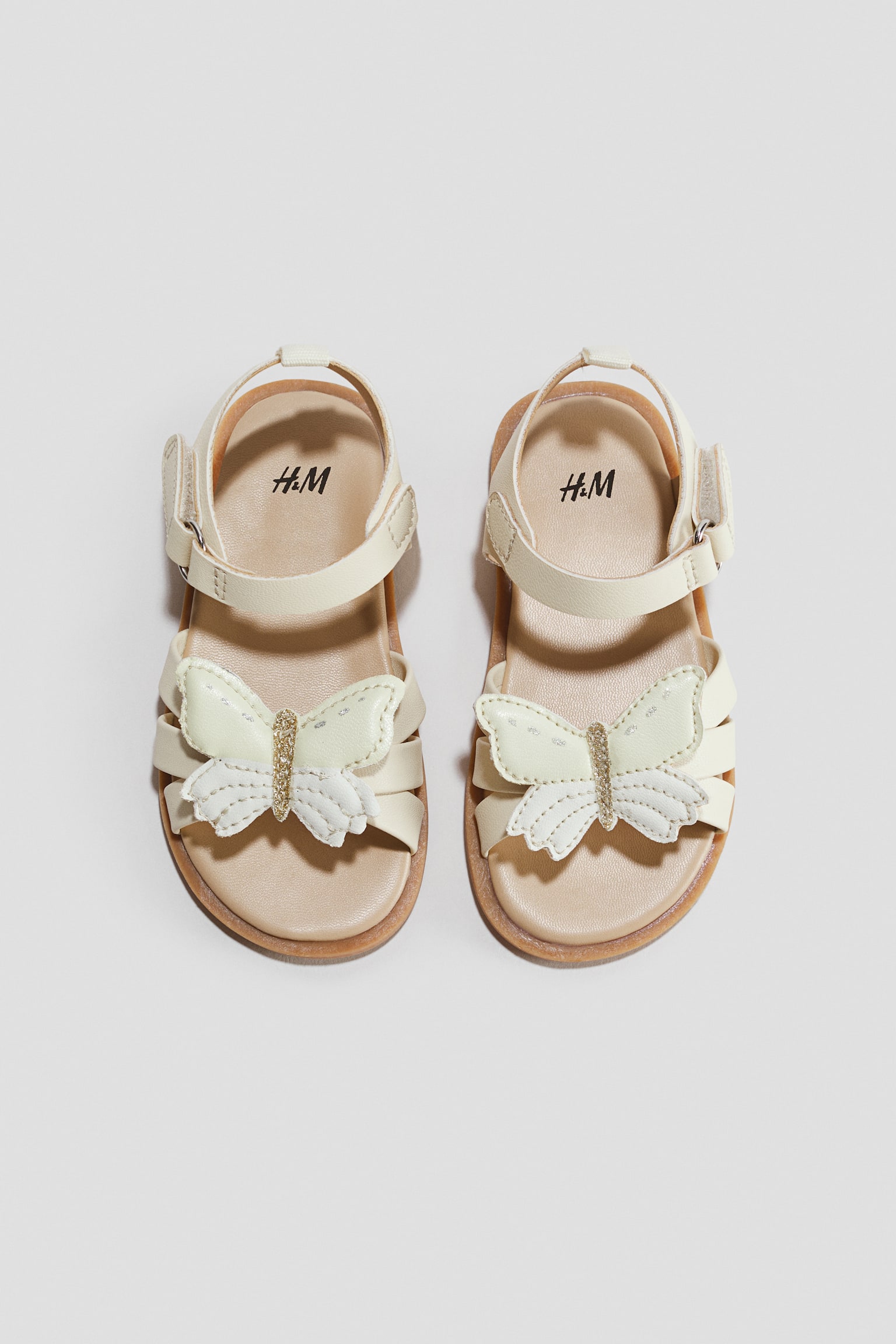 Strappy sandals in cream-white are shown facing front in a top-down view, showcasing a large, layered butterfly applique with a glittery gold body over the toe straps. They feature adjustable ankle straps with hook-and-loop fasteners, light tan insoles, and thin brown outsoles.