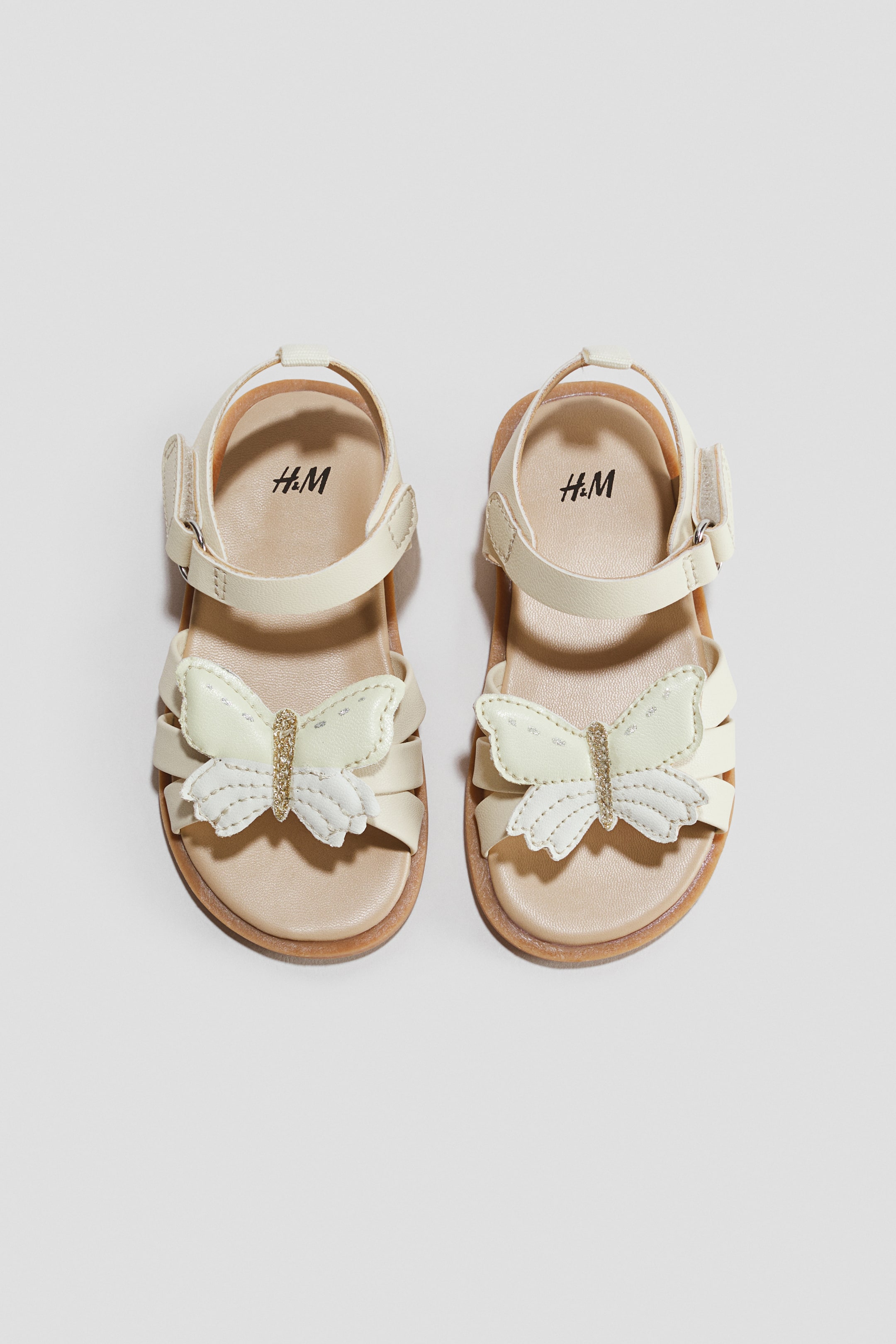 View larger image: Strappy sandals in cream-white are shown facing front in a top-down view, showcasing a large, layered butterfly applique with a glittery gold body over the toe straps. They feature adjustable ankle straps with hook-and-loop fasteners, light tan insoles, and thin brown outsoles.