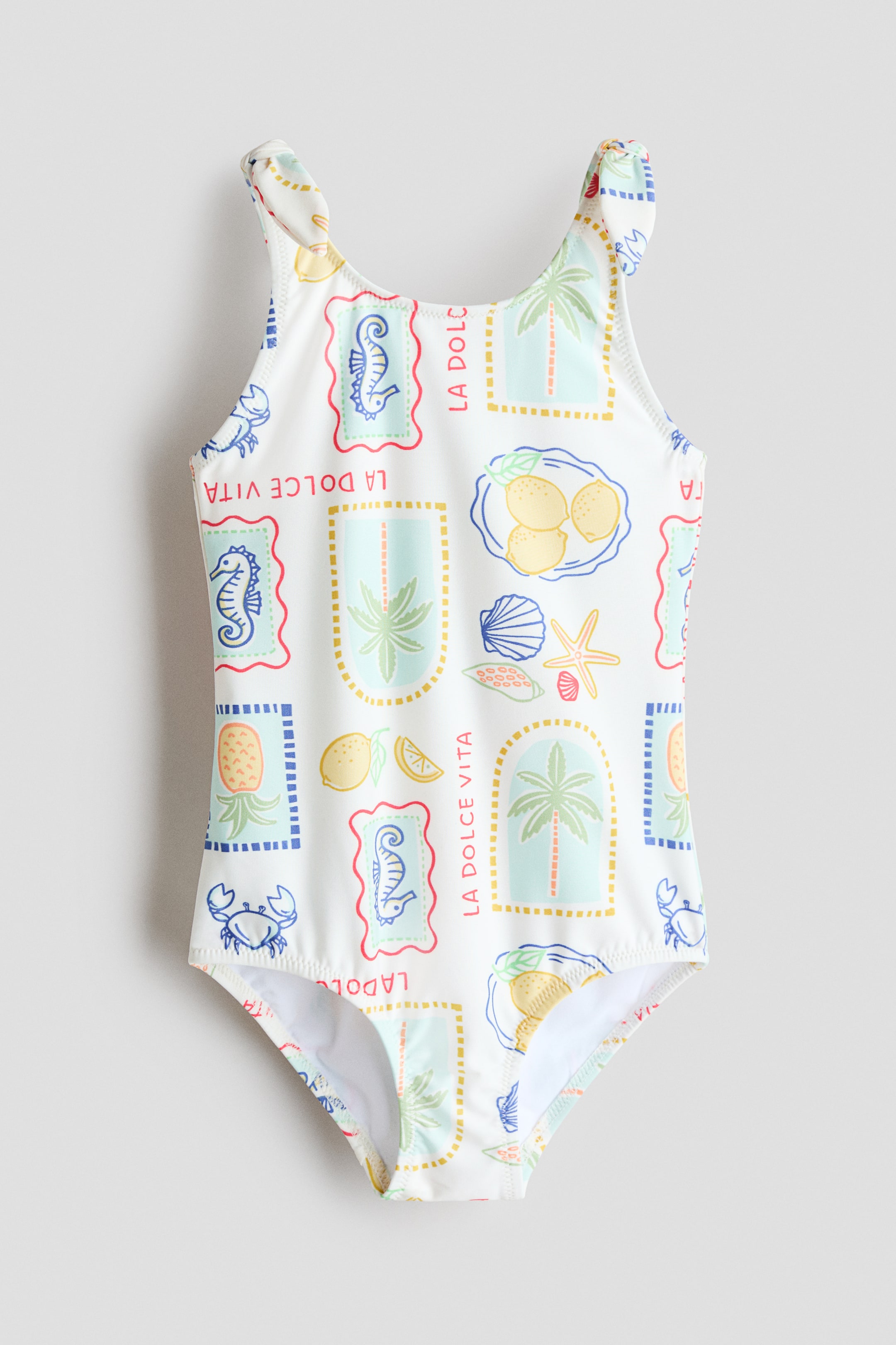 View larger image: The swimsuit showcases its front, adorned with an all-over print of stylized stamps featuring a variety of summer motifs such as lemons, palm trees, seahorses, and the text 'LA DOLCE VITA'. It highlights shoulder straps accented with decorative knot details.