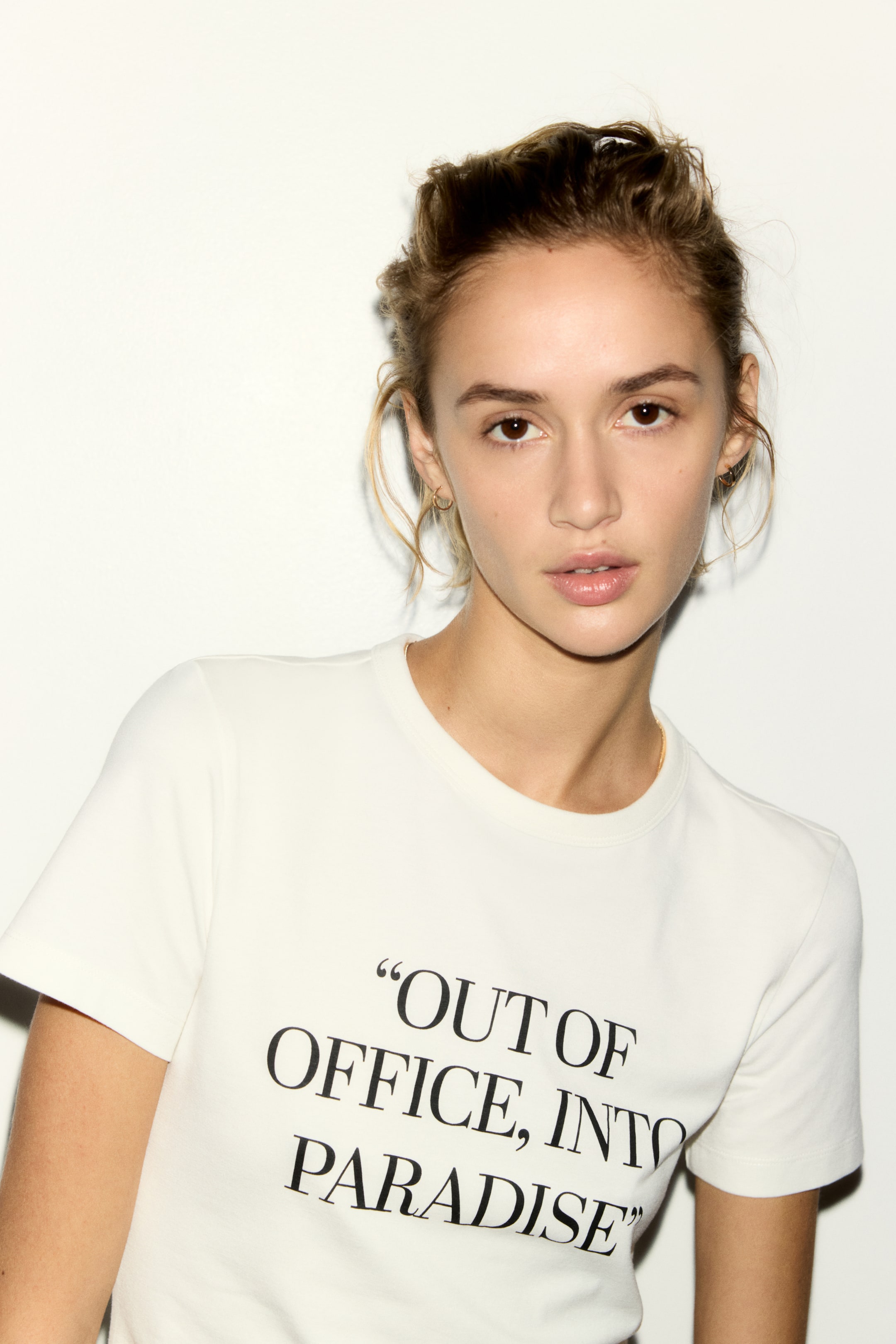 Se større billede: Modellen bærer en off-white tætsiddende t-shirt. Den har et sort printmotiv på forsiden med teksten 'OUT OF OFFICE, INTO PARADISE', en rund halsudskæring med ribkant og korte ærmer.