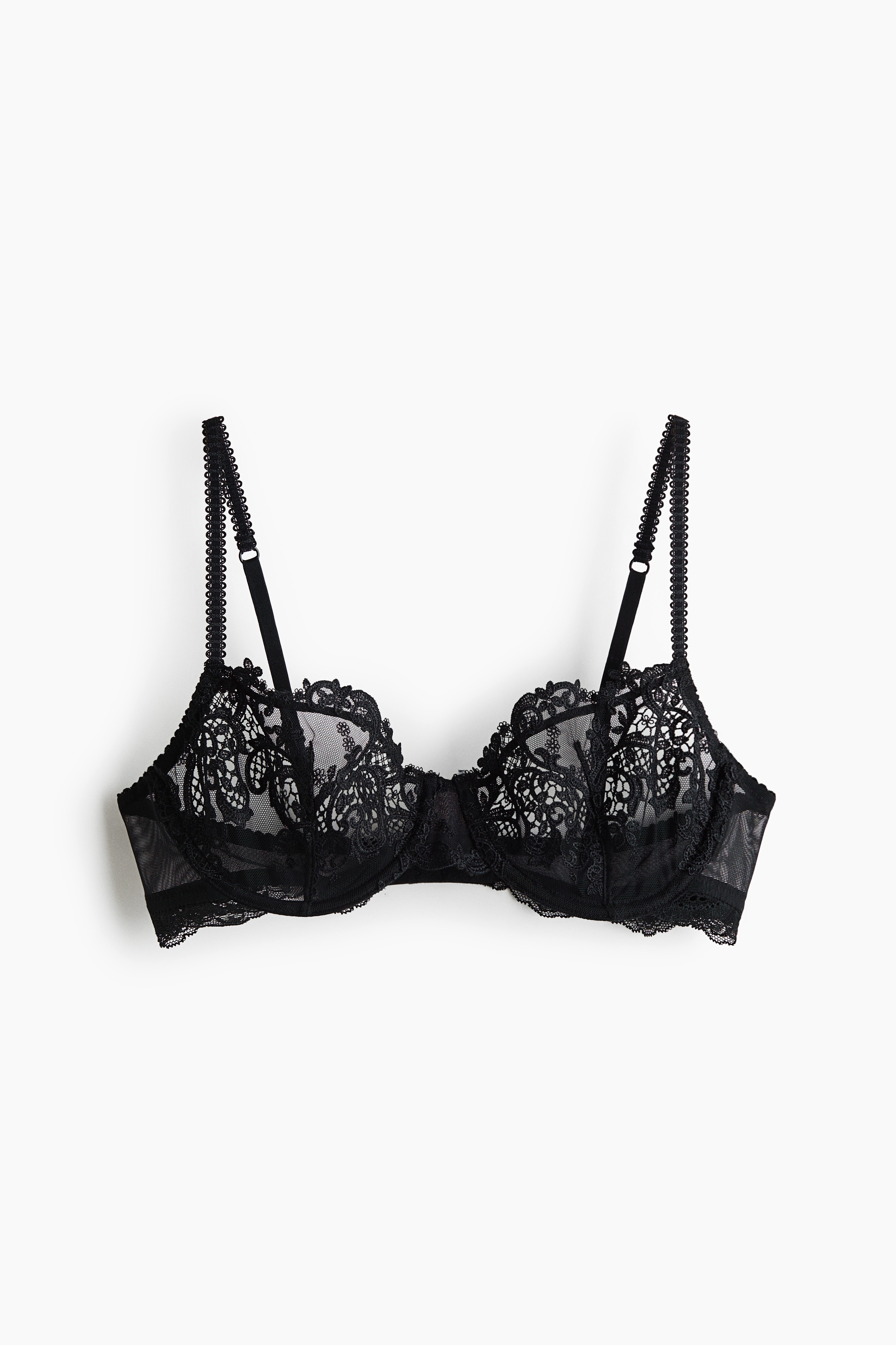 Una modella indossa un reggiseno nero con ferretto in pizzo, caratterizzato da coppe trasparenti con ricami floreali intricati e sottili spalline smerlate. Il reggiseno presenta una silhouette scollata che mette in risalto i delicati dettagli in pizzo semi-trasparente.