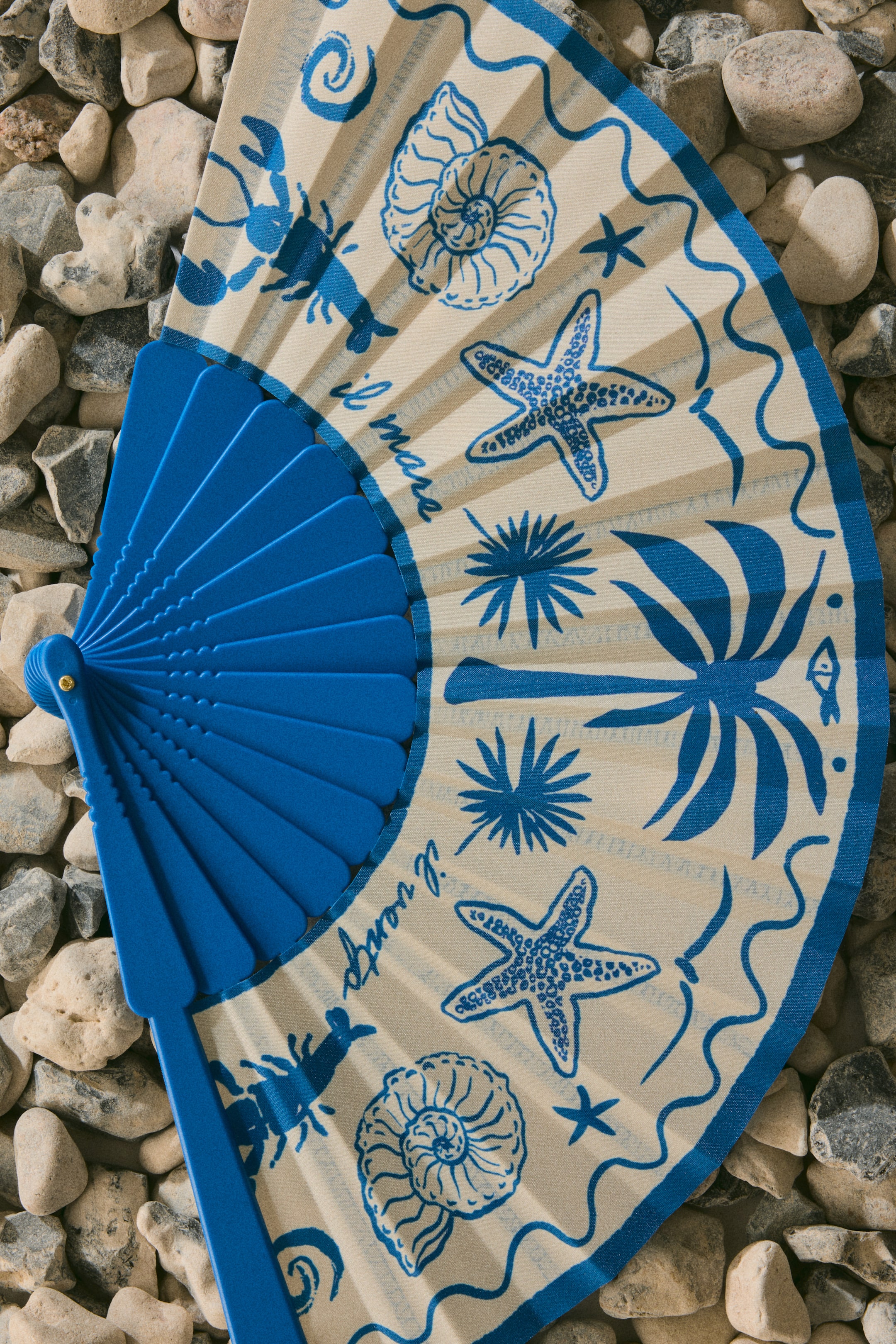 View larger image: A folding fan showcases a cream-hued fabric leaf adorned with blue illustrations of starfish, shells, lobsters, palm-like plants, and blue script text reading "il mare" and "il vento." This elegant design fans out from a solid blue ribbed handle, featuring a blue border along its outer edge.