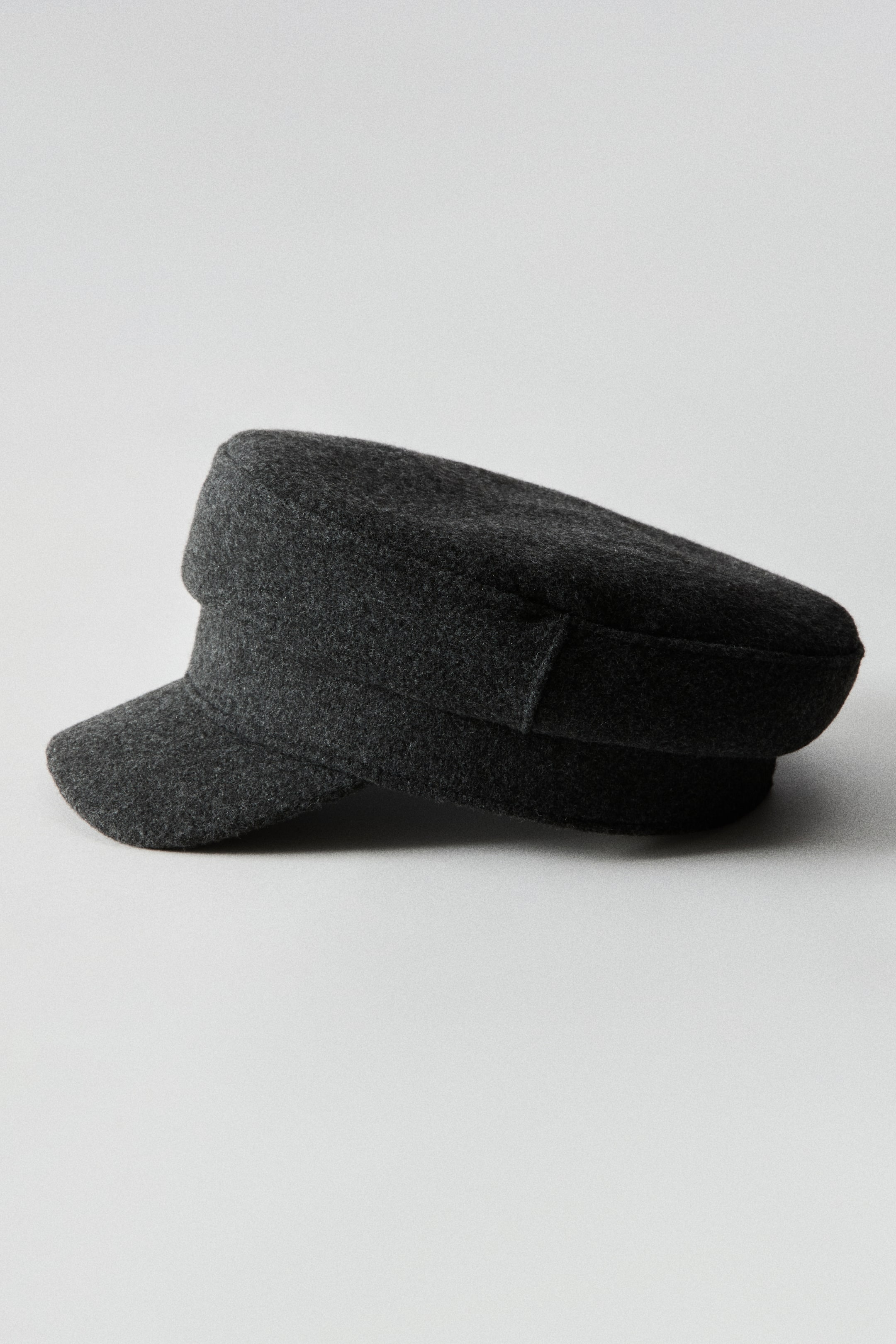 Ampliar la imagen: Se muestra una gorra baker boy en tela tejida texturizada de color gris oscuro, en vista lateral orientada hacia la izquierda. La gorra presenta una copa estructurada con parte superior plana, una banda ancha que rodea la base y una visera corta y curva.