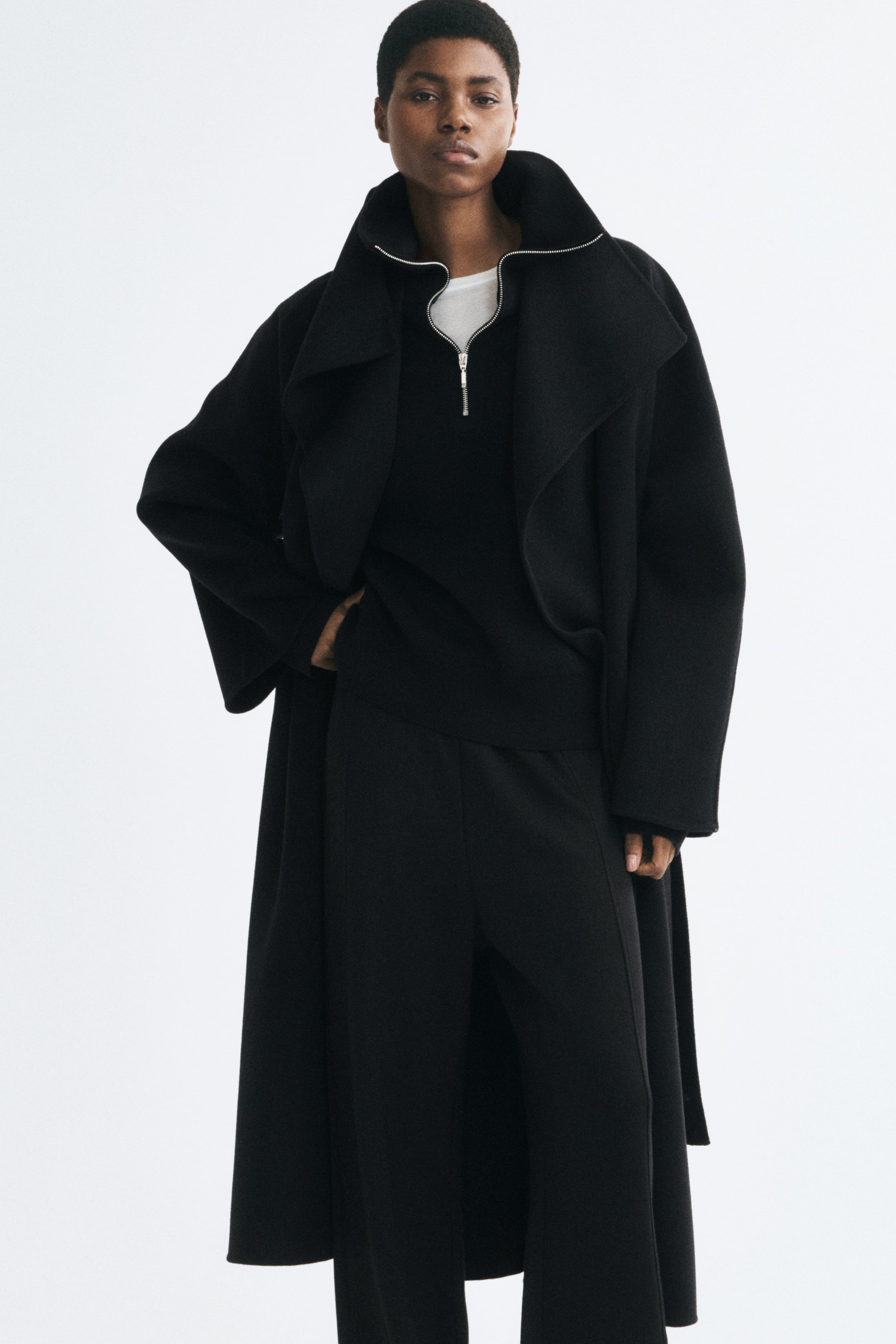 View larger image: A black fine-knit sweater featuring a ribbed funnel neck with a partially open silver half-zip is worn by the model. The sweater is styled in layers under a long, heavyweight black coat and over a white T-shirt, paired with matching black trousers.
