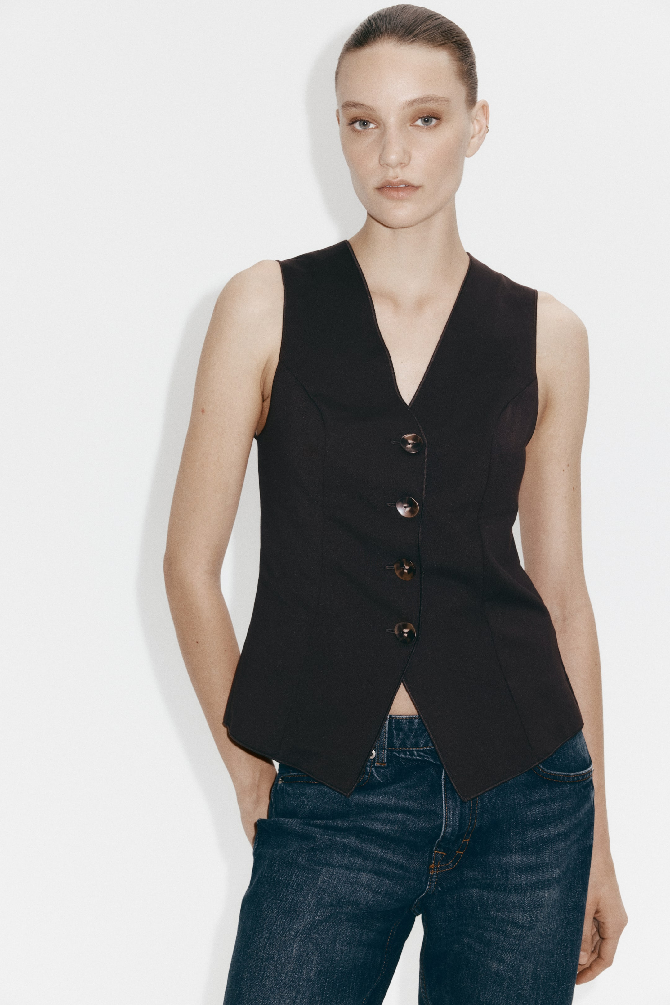 Visualizza immagine più grande: Gilet senza maniche di colore scuro con scollo a V, chiusura frontale a quattro bottoni con l’ultimo lasciato sbottonato, cuciture sartoriali e orlo frontale a punta. Abbinato a jeans in denim scuro.