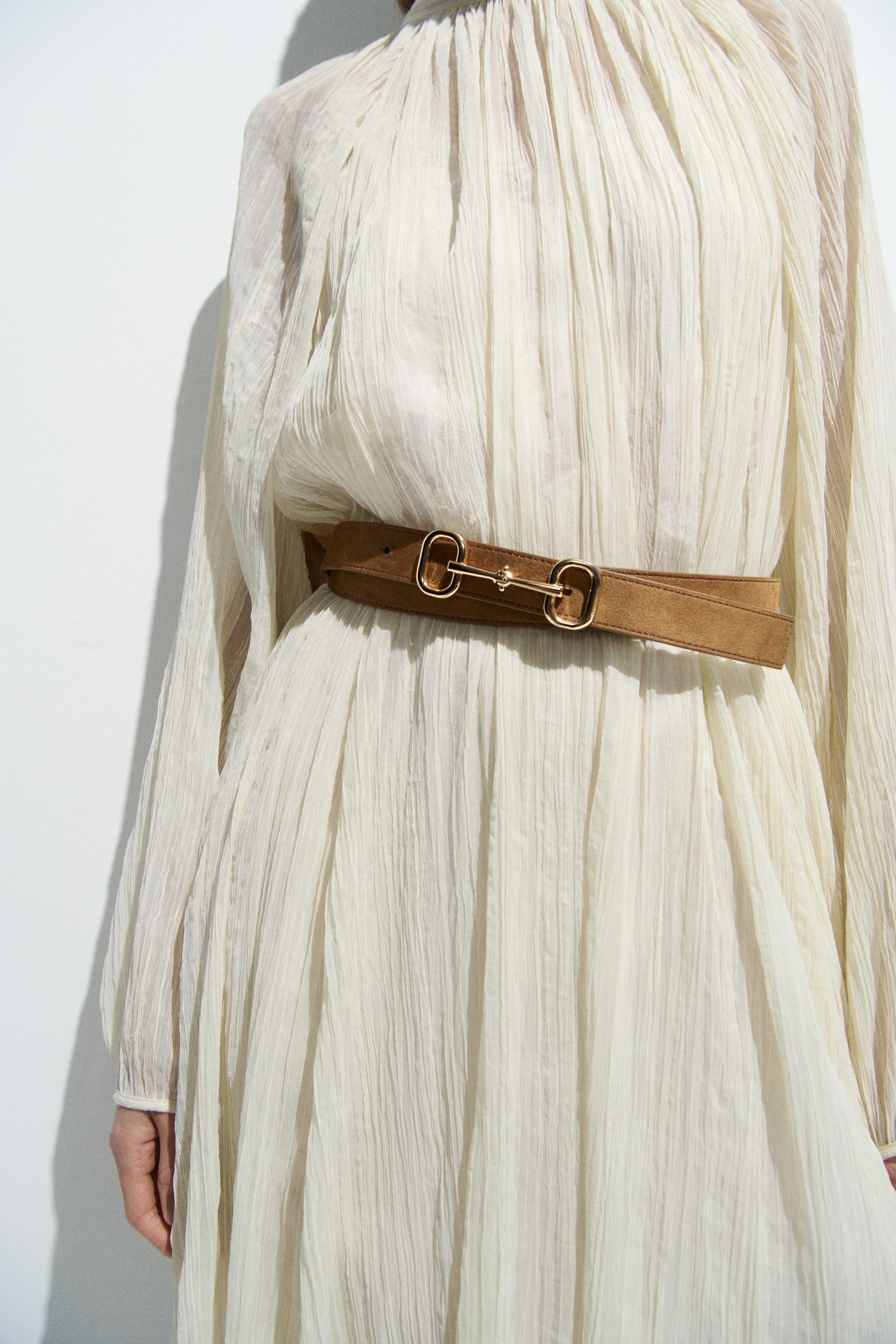 View larger image: A tan suede belt is wrapped in a double layer around the model's waist. It fastens with a gold-toned metal snaffle buckle, cinching a cream pleated dress.