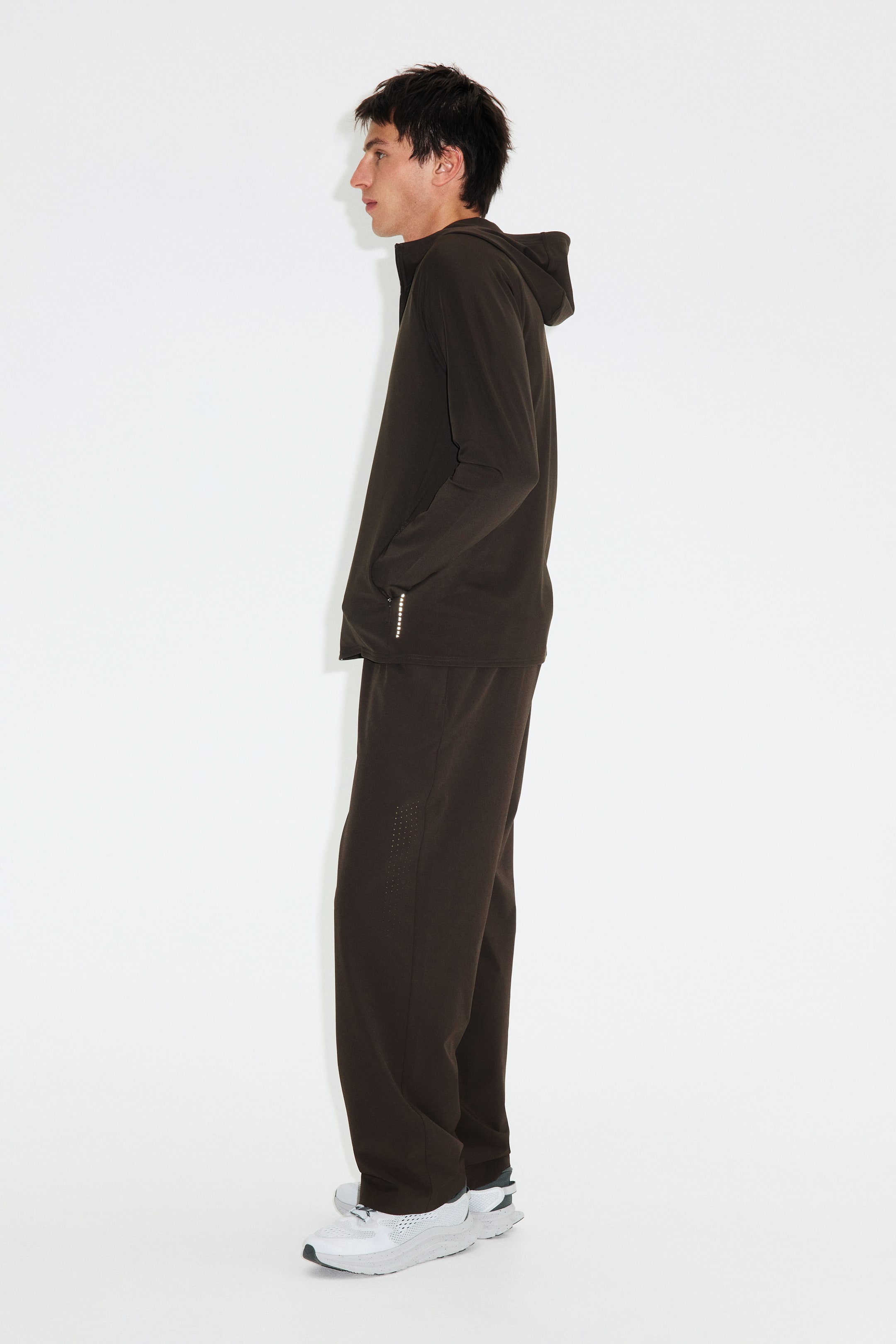 Zobacz większy obraz: Ciemnobrązowa bluza z kapturem zapinana na zamek oraz pasujące spodnie dresowe prezentowane są z boku. Bluza posiada kaptur, długie rękawy oraz odblaskowy detal na lewym mankiecie, podczas gdy spodnie wyróżniają się perforowanymi detalami na górnej części lewego uda.