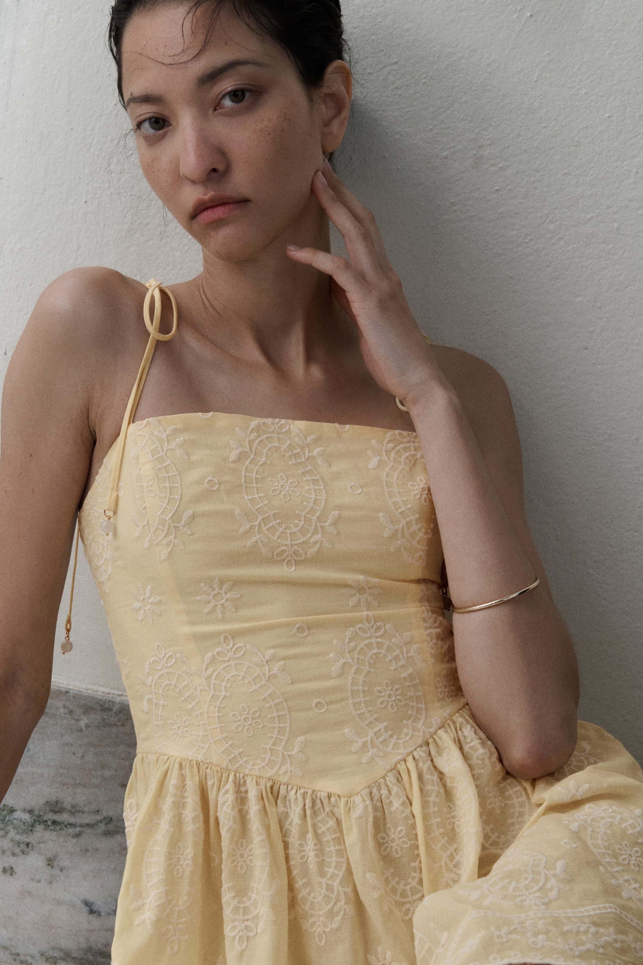 Voir une image plus grande: Une robe courte en tissage de coton jaune pâle avec une broderie blanche sur toute la surface est portée par le mannequin. Elle présente une coupe droite en haut et des bretelles spaghetti à nouer avec de petites perles décoratives. La robe est dotée d'un corsage ajusté avec des coutures princesse, d'une couture en V froncée au-dessus des hanches et d'une jupe évasée.