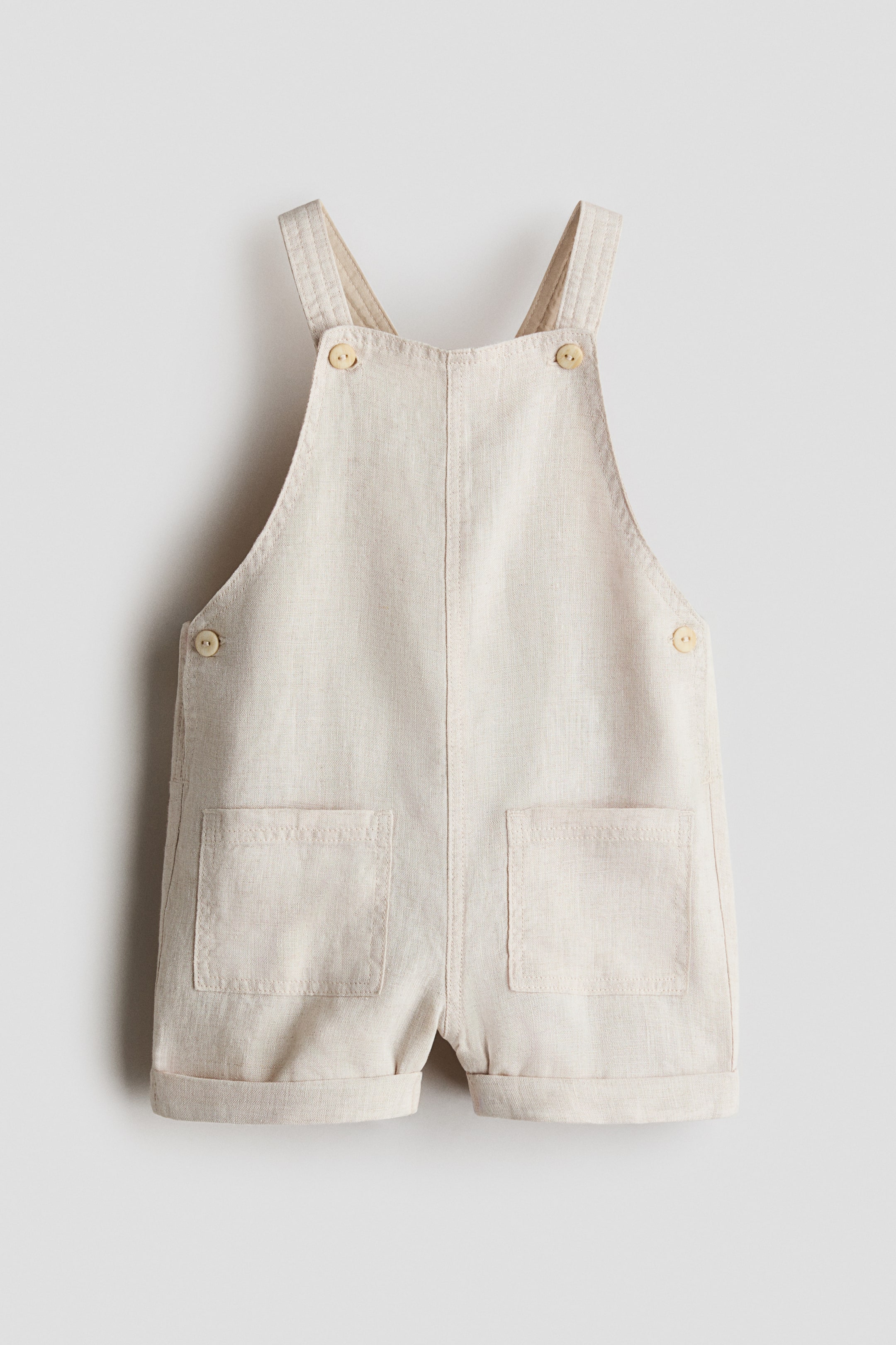 Grösseres Bild ansehen: Kinderkurze Overalls präsentieren sich frontseitig aus einem hellbeigen, strukturierten Stoff. Sie verfügen über zwei Schulterträger mit hellen Knöpfen am Latz, zwei Seitenknöpfe, zwei aufgesetzte Vordertaschen und hochgekrempelte Säume an den Shorts.