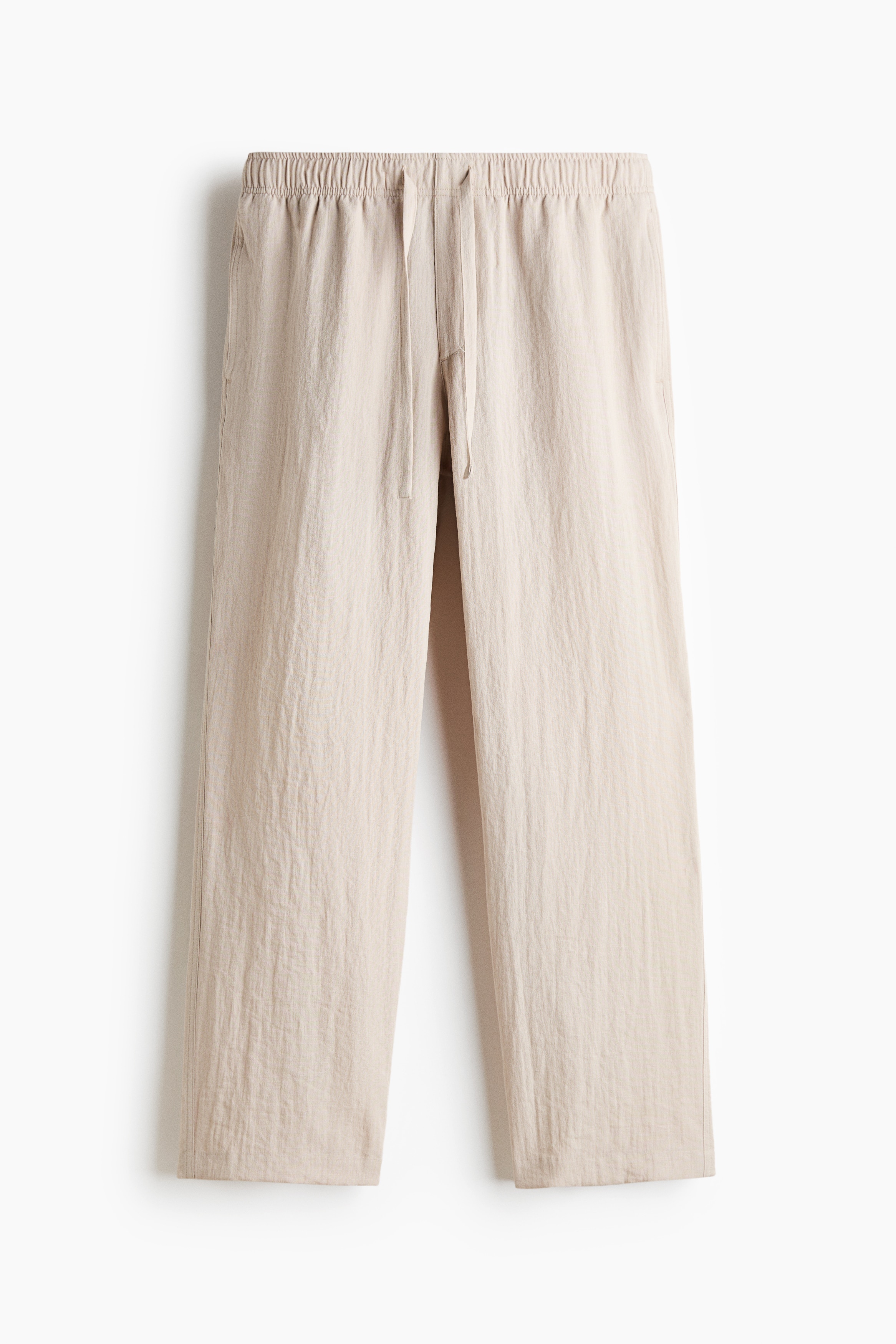 Straight-leg trousers in a light beige crinkled-texture weave are worn by the model, featuring a relaxed fit, an elasticated waistband and a drawstring. The model is standing with one hand in a side pocket, styling the trousers with a short-sleeved dark grey button-up shirt and light-coloured trainers.