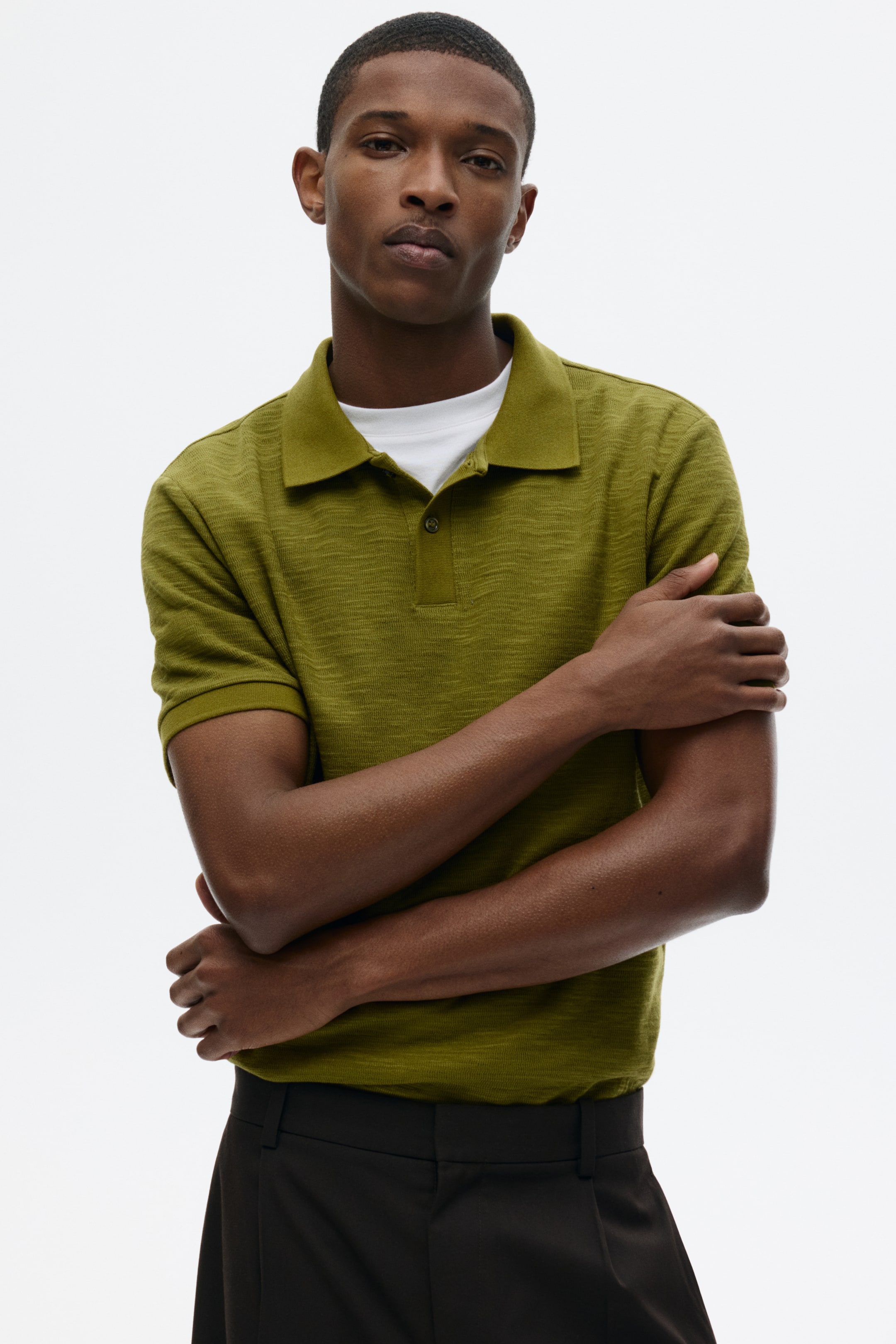View larger image: An olive-green textured knit polo shirt showcases its classic collar and a two-button placket. It features short sleeves and is layered over a white T-shirt, which is visible at the neckline.