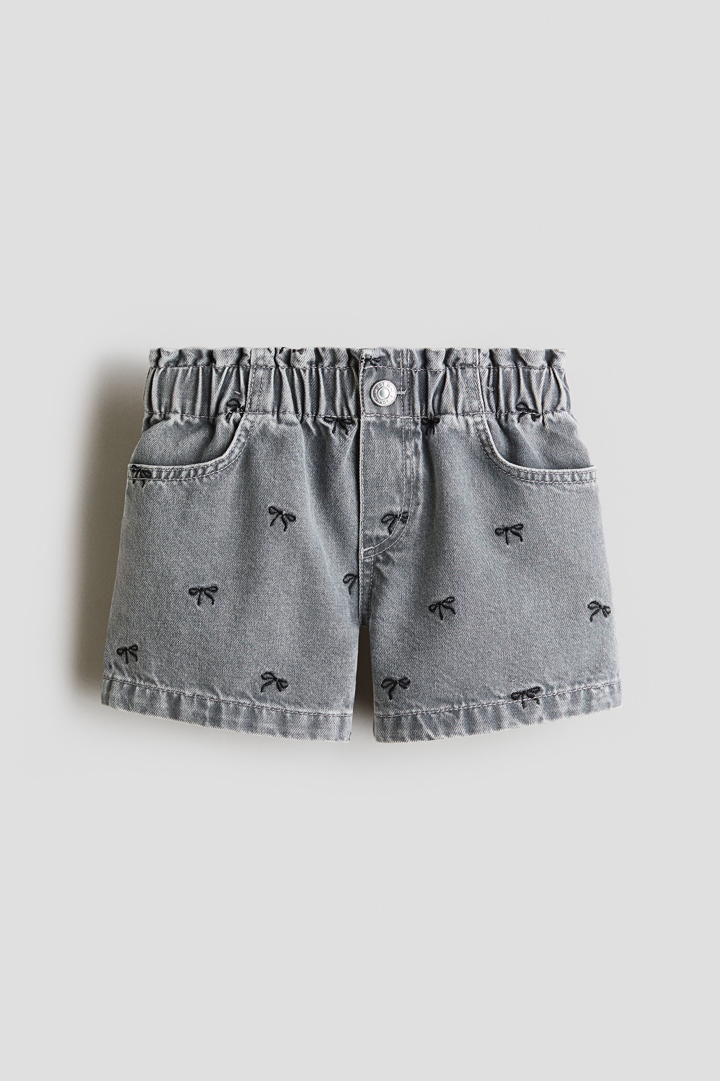 These grey cotton-weave shorts are shown from the front, featuring an elasticated paperbag waist, a mock fly with a press stud, and scattered black embroidered bows. A cream and black striped long-sleeve top is being worn tucked into the waistband, accentuating the paperbag style and front pockets.