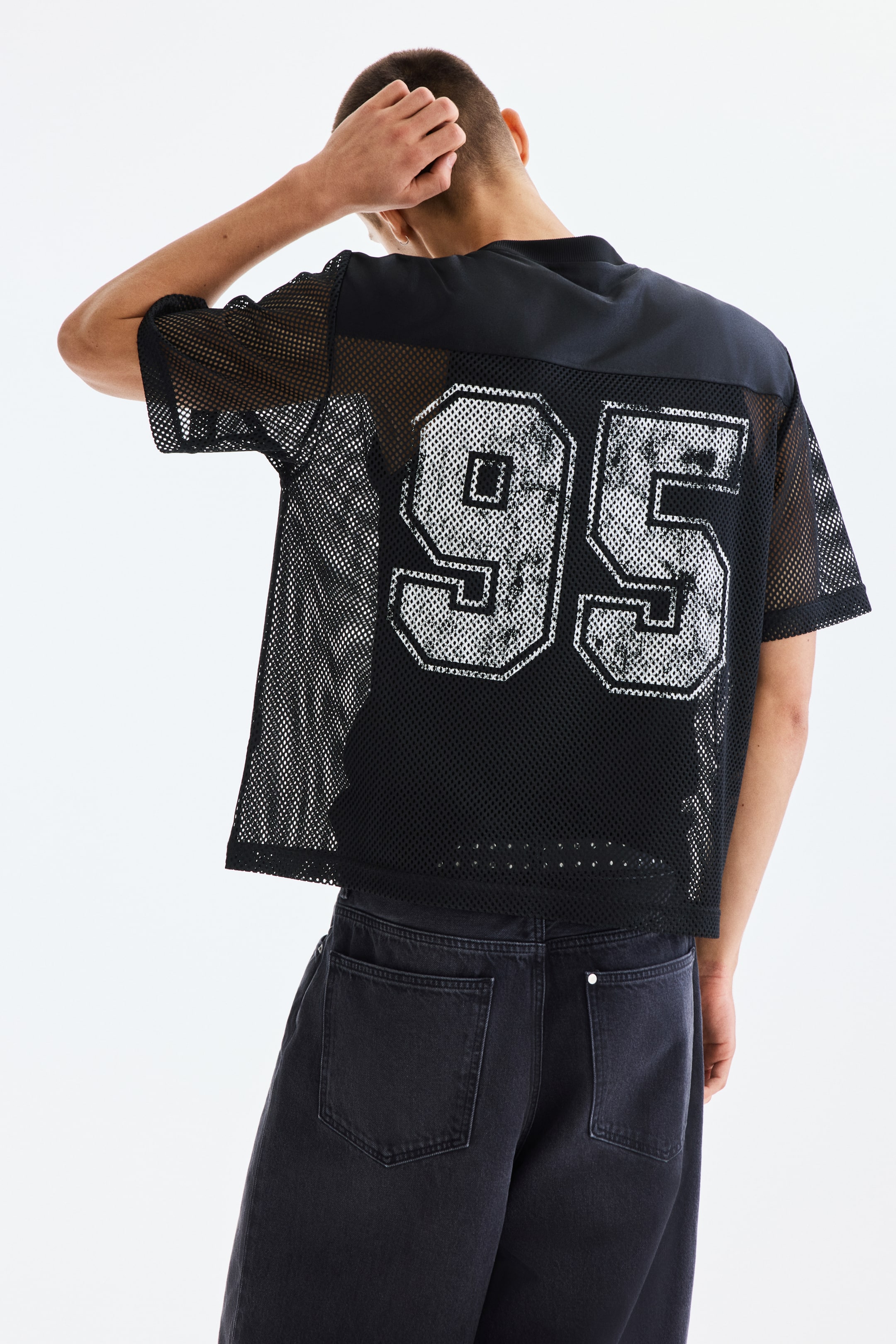 View larger image: A black mesh jersey is being viewed from the back, featuring short sleeves and solid black fabric across the shoulders and upper back. A large white distressed number "95" with black outlines is centered on the back, and the jersey is being worn over a black t-shirt.