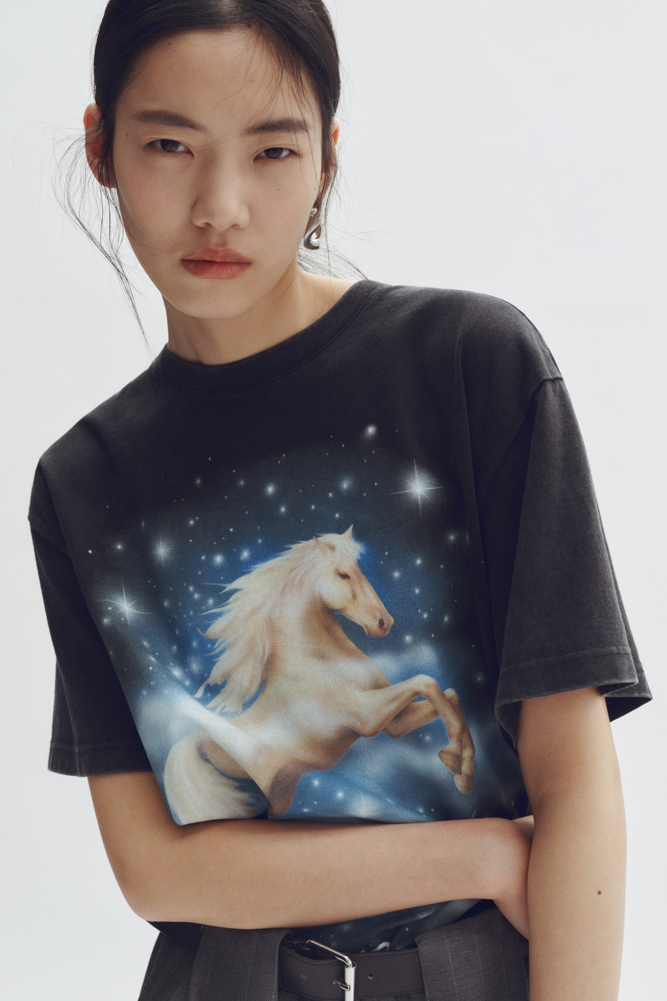 Grotere afbeelding bekijken: Een donkergrijs T-shirt met een grote afbeelding van een lichtgekleurde, steigerende paard tegen een sterrenrijke blauwe kosmische achtergrond. Het is ontworpen met een geribbelde ronde hals en korte, relaxed-fit mouwen.