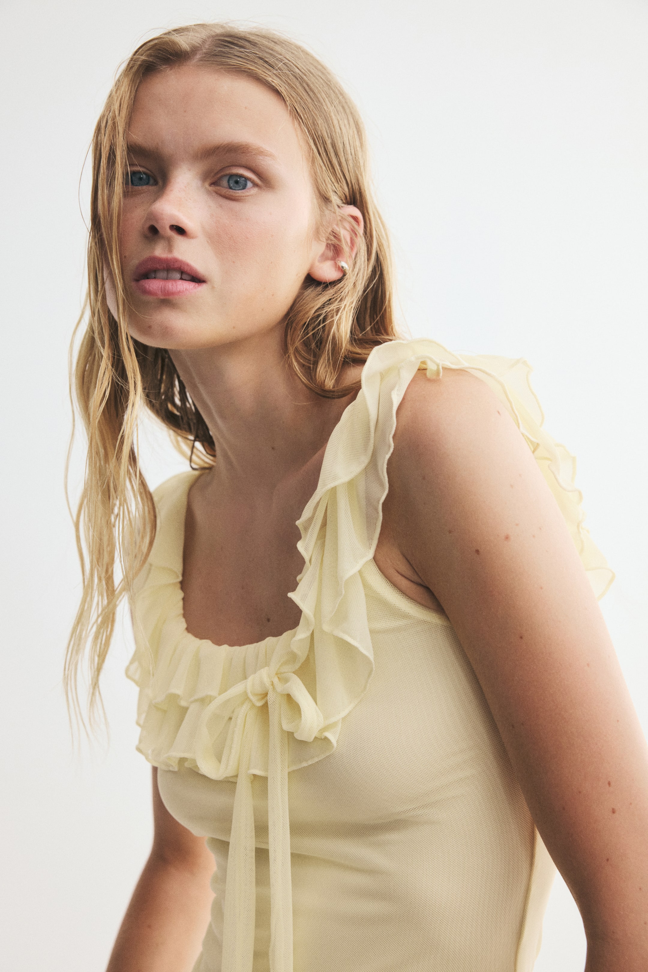 View larger image: A pale yellow fitted top in sheer mesh is being worn by the model. It features a scoop neckline with tiered frill trims extending over the shoulders, wide shoulder straps, and a decorative tie detail at the front, with the mesh layered over a solid lining.