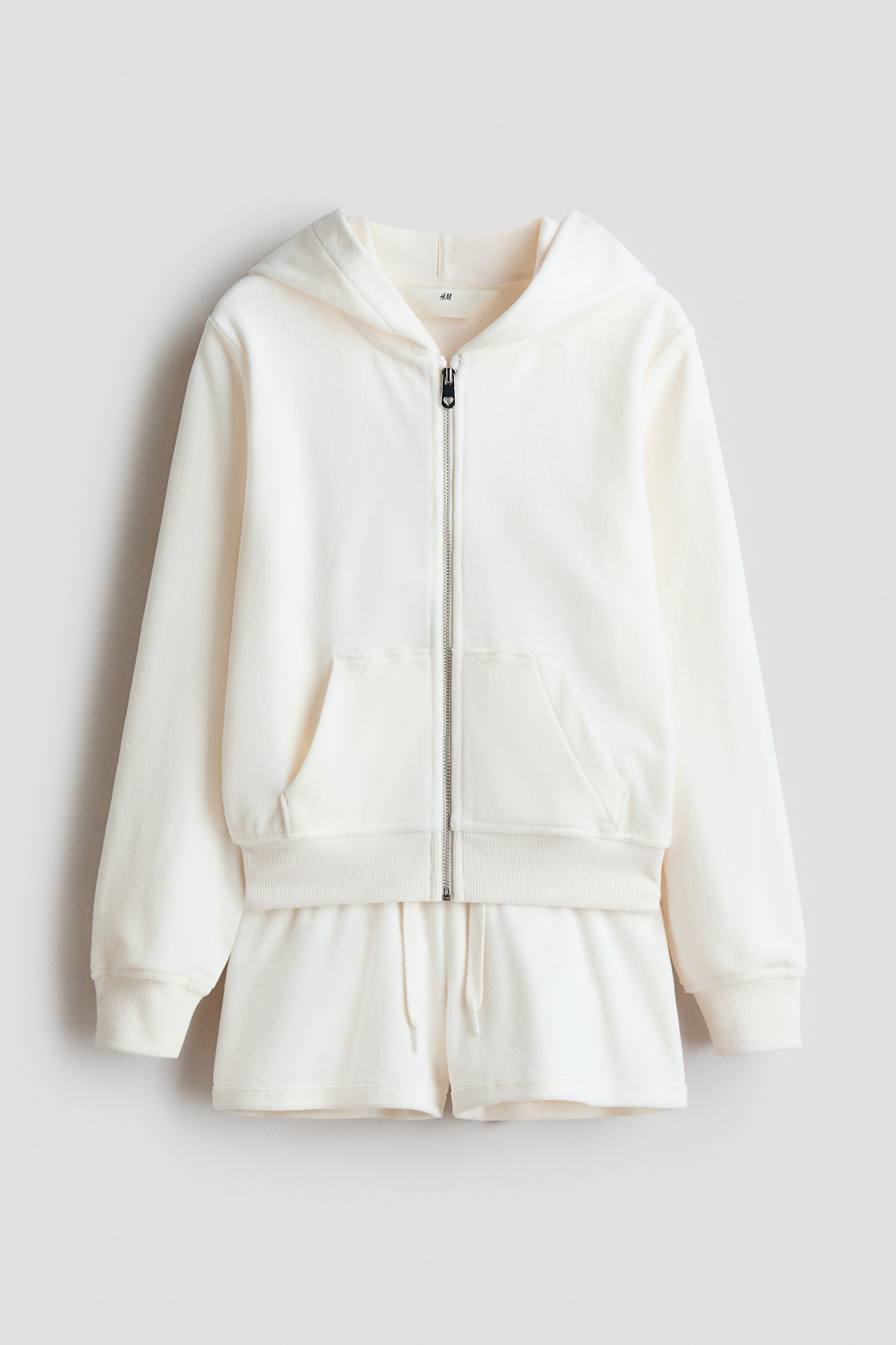 View larger image: A cream-coloured loungewear set in plush fabric is shown from the front, consisting of a full-zip hoodie and matching shorts. The hoodie features a hood, a silver-tone zip with a heart-shaped pull tab, a split kangaroo pocket, and ribbed cuffs and hem. The shorts feature a drawstring waist.
