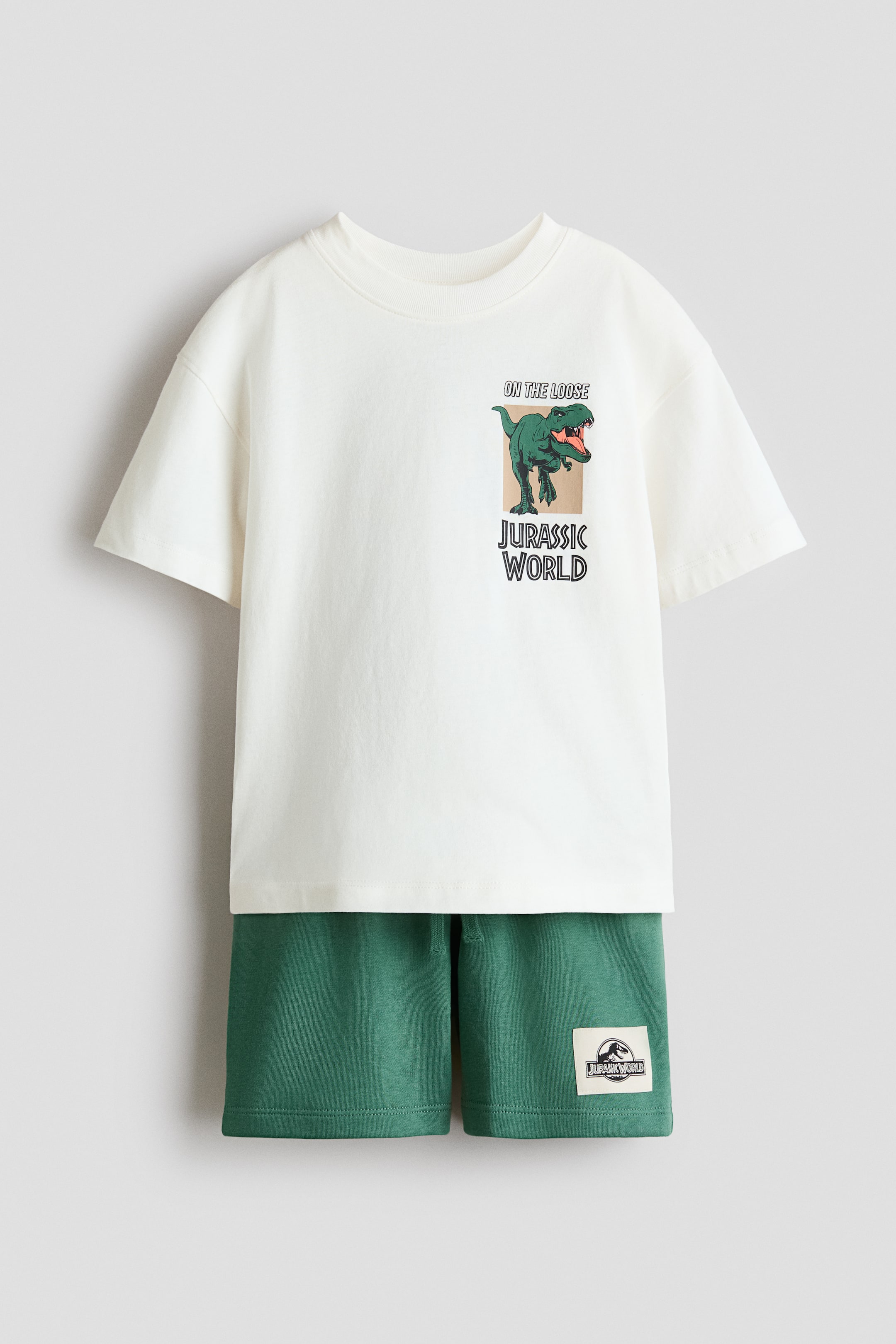 Voir une image plus grande: Un ensemble assorti face avant comprend un T-shirt en jersey de coton blanc cassé avec des épaules tombantes, des manches larges et un col côtelé, accompagné de shorts vert foncé. Le T-shirt arbore une impression graphique sur la poitrine gauche d'un T-Rex vert avec les textes « EN CAVALE » et « JURASSIC WORLD », tandis que les shorts sont confectionnés en tissu de sweat léger et incluent une taille à cordon de serrage avec un écusson du logo « Jurassic World » sur la jambe droite.