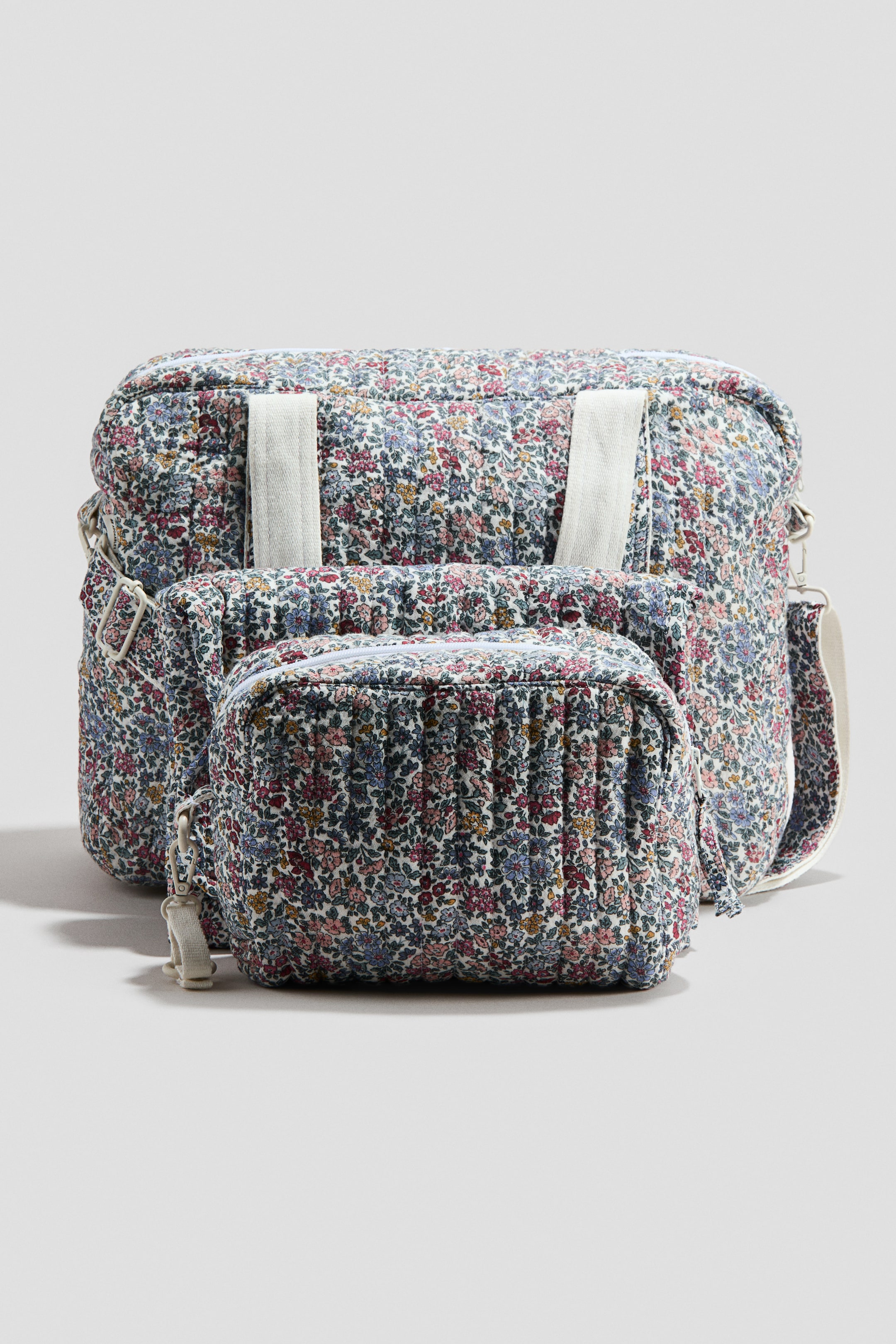 Prikaži veću sliku: Set podloženih torbi za presvlačenje prikazan je s prednje strane, uključujući veliku torbu za pelene i manju torbu za pranje, obe izrađene od prošivenog pamučnog muslina i sa cvetnim printom u ružičastim, plavim, zelenim i žutim nijansama. Torba za pelene ima dva bela platnena ručka na vrhu i podesivi remen za rame od belog platna sa karabin kukom sa strane.