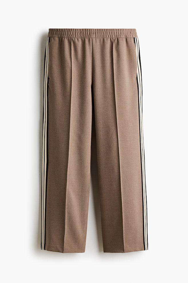 The brown pants are displaying an elasticized waistband and a vertical crease running down the front of each leg. Triple stripes, consisting of black and light yellow bands, are accentuating the outer seams of both pant legs. - 1