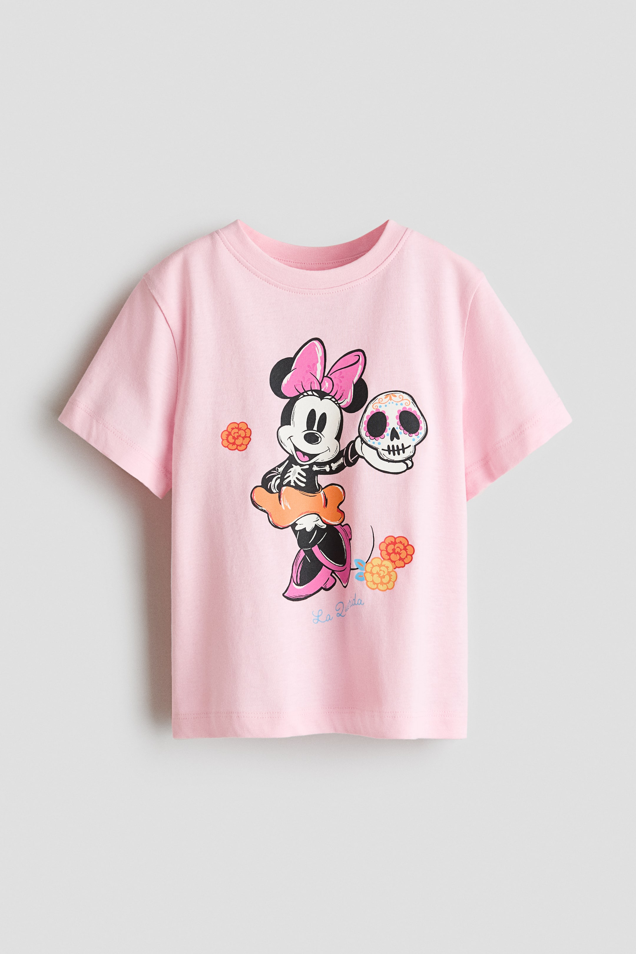 View larger image: A pink short-sleeve T-shirt features a crew neck. The front displays a graphic of Minnie Mouse dressed as a skeleton, wearing a pink bow and orange skirt, and holding a decorated sugar skull, with small floral elements and the text 'La Vida'.