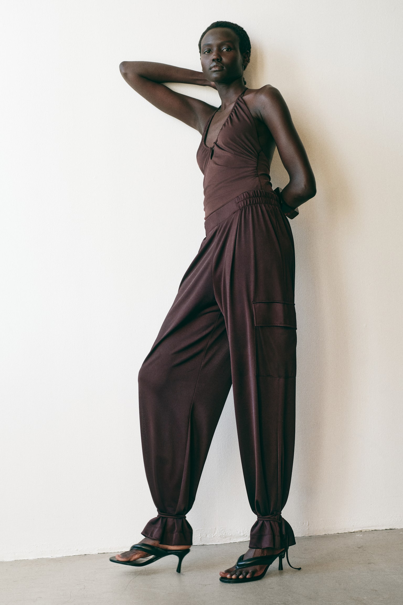 Oversized dark brown jersey trousers are showcased from the front, featuring a regular waist with covered elastication, balloon legs with flap pockets, and drawstrings cinching the flounced hems at the ankles. - 1