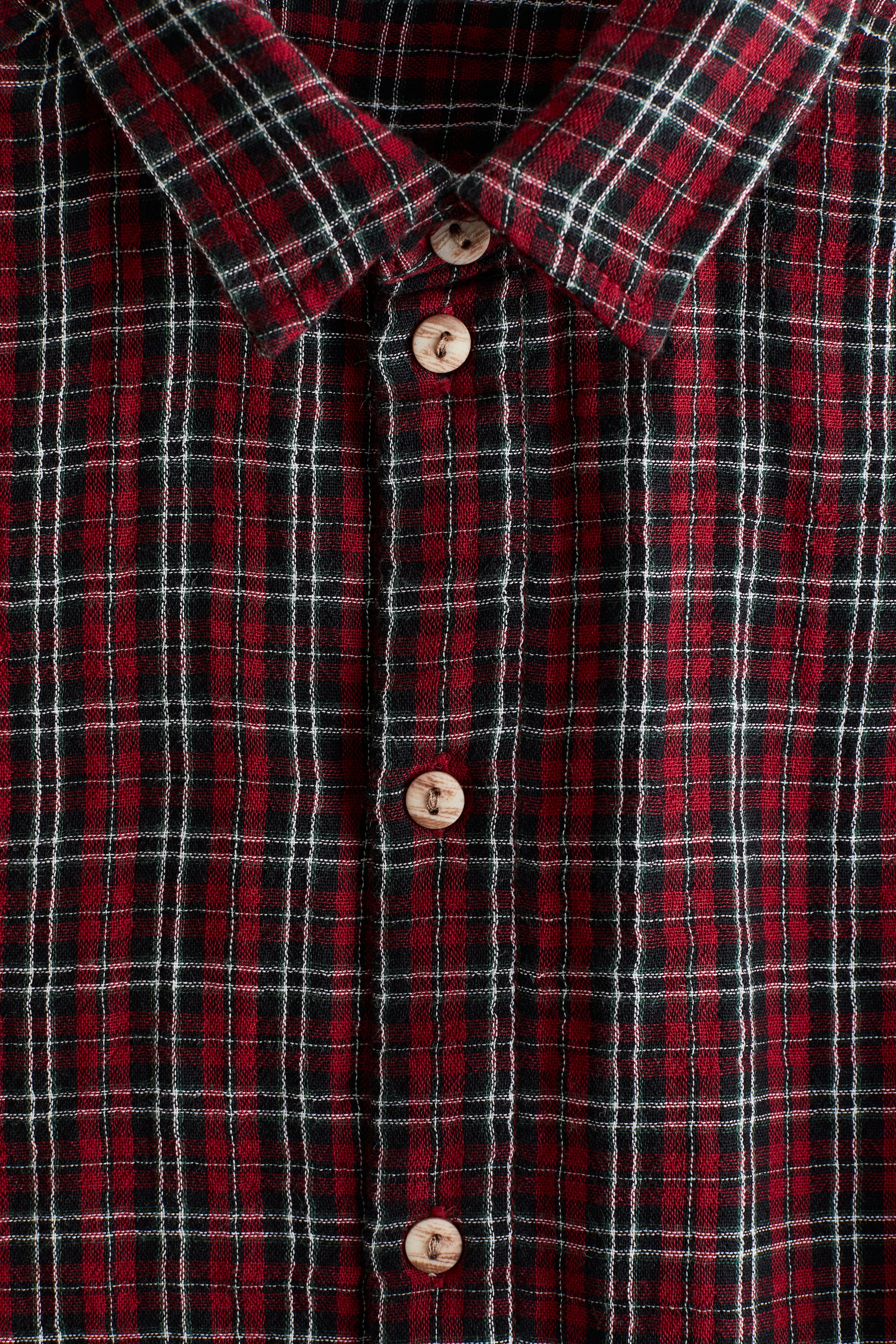 Visualizza immagine più grande: Una camicia a quadri rossa e nera è presentata dal davanti, caratterizzata da un tessuto intrecciato con motivo a quadri rosso, nero e bianco. È visibile un colletto a punta e una lista di bottoni centrale è chiusa con bottoni rotondi in legno di colore chiaro.