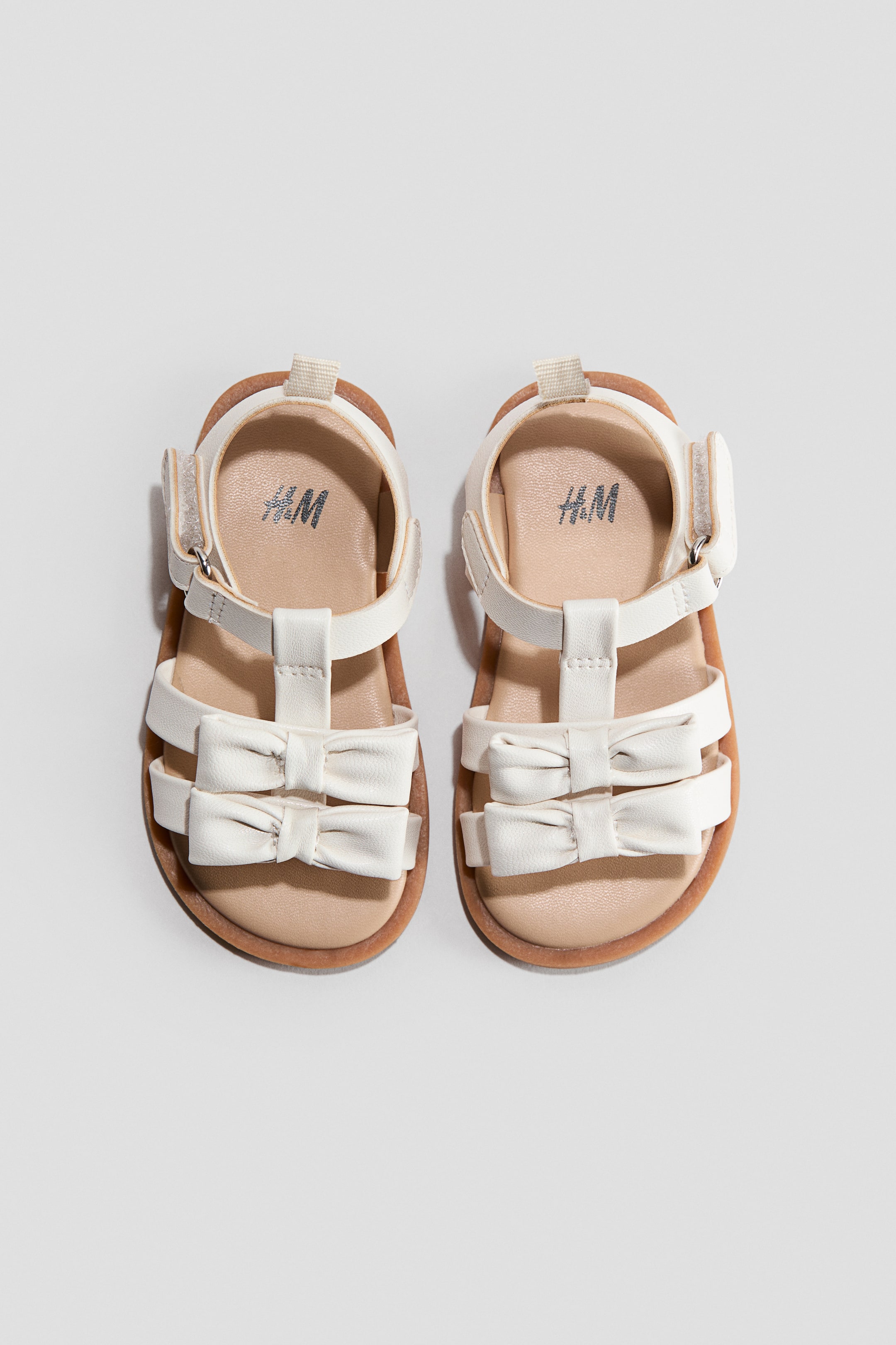 Xem ảnh lớn hơn: Một đôi dép sandal màu kem dành cho trẻ em có thiết kế quai chữ T và hai quai ngang ở phần trước bàn chân, mỗi quai được trang trí bằng một chiếc nơ nhỏ. Quai cổ chân có khóa dán hook-and-loop có thể nhìn thấy, cùng với đế chân màu be nhạt nằm trên đế nâu, hiển thị logo H&M.