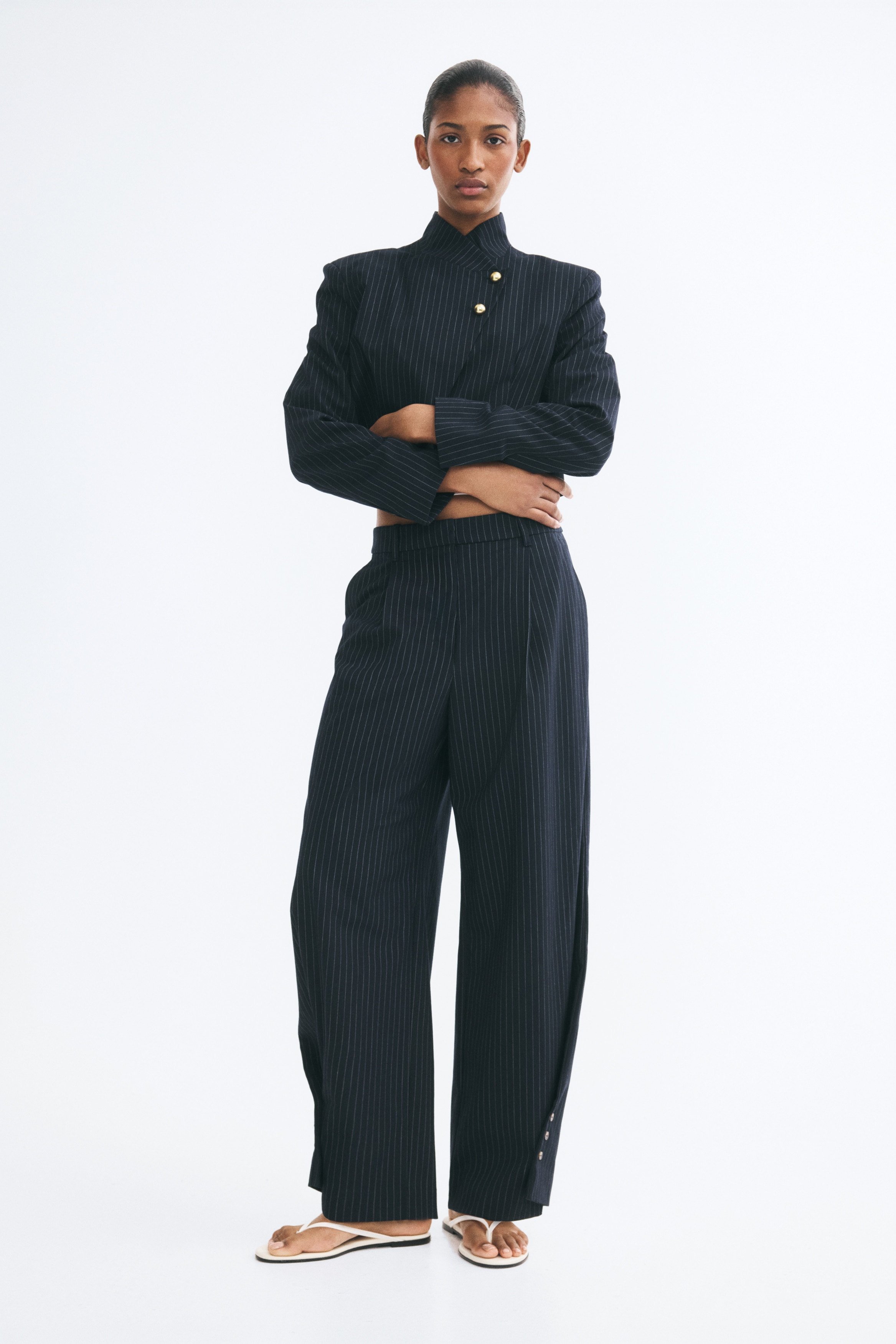 A pair of navy blue, pinstriped woven pants is hanging facing front, displaying a high waist with front pleats. The barrel-leg silhouette tapers towards the bottom, showcasing unique wrap-around sides secured with buttons at the outer hems.
