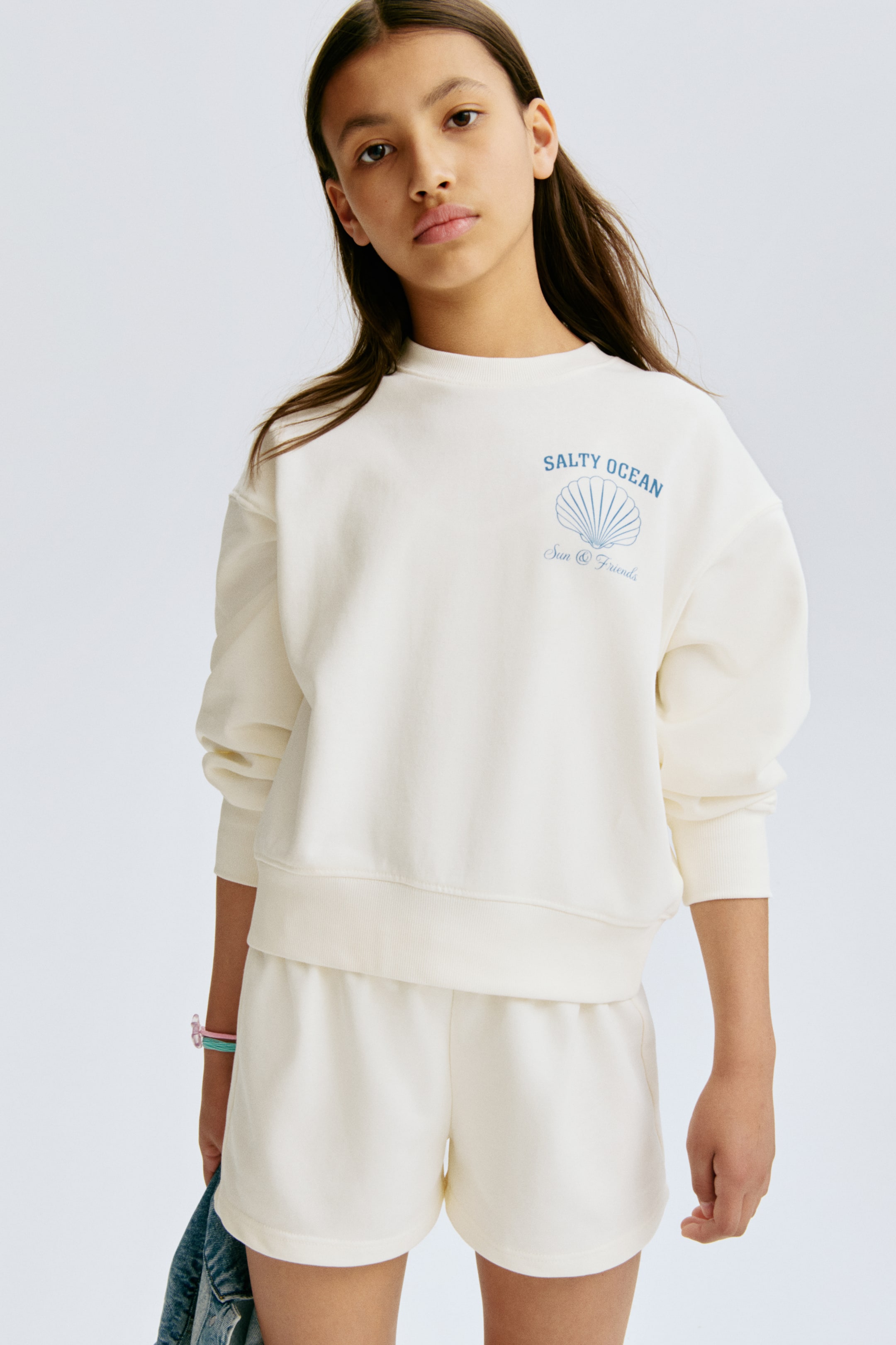 Visualizza l'immagine ingrandita: Una felpa color crema e pantaloncini coordinati vengono presentati frontalmente. La felpa presenta il testo blu "SALTY OCEAN" e un grafico di conchiglia sul petto sinistro, insieme a polsini e orlo a coste. I pantaloncini coordinati mostrano una vestibilità rilassata e una cintura arricciata.