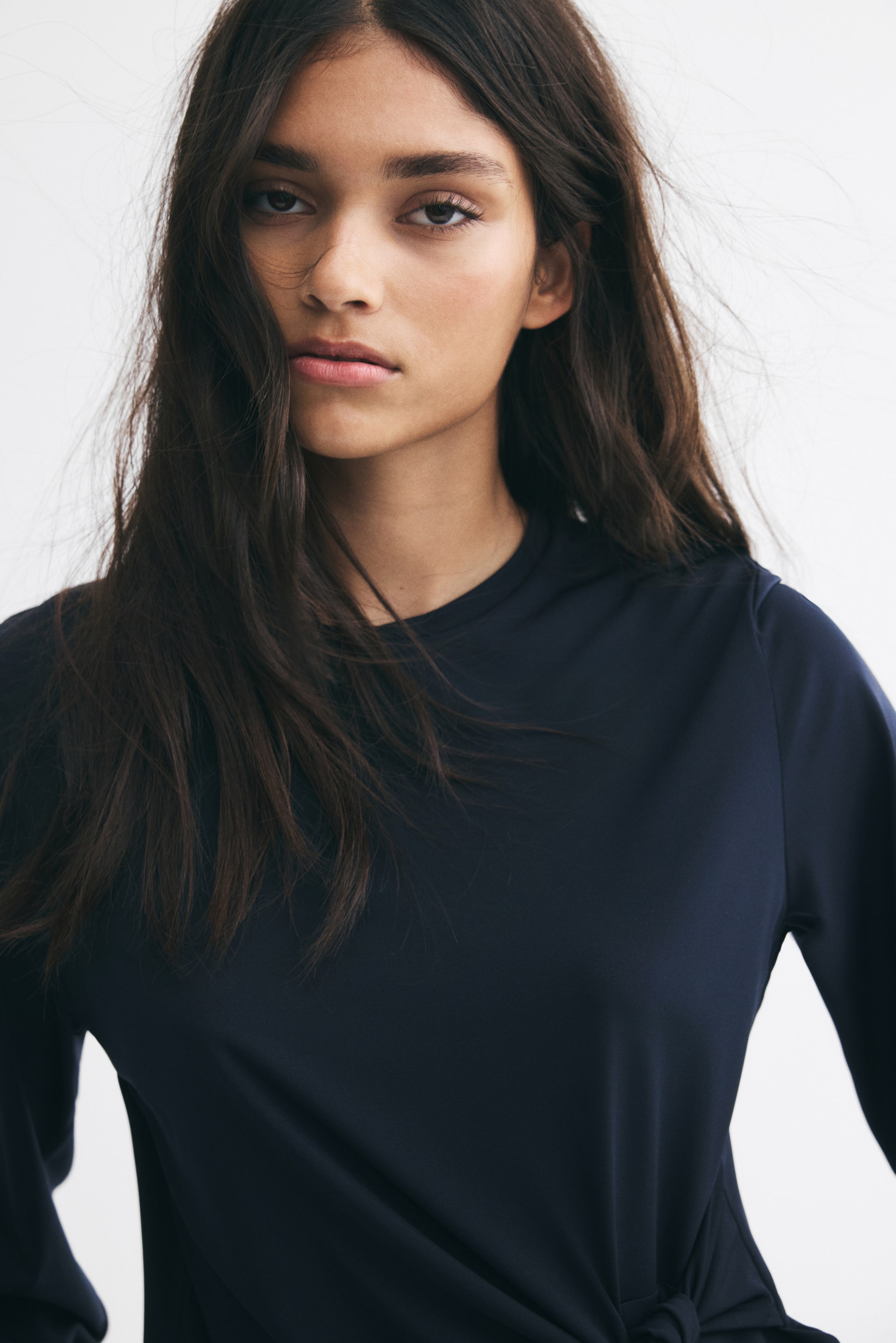 View larger image: A dark navy long-sleeve top made of smooth stretch jersey is worn by the model. The top features a round neckline and a knotted tie detail at one side of the waist, creating a draped effect.