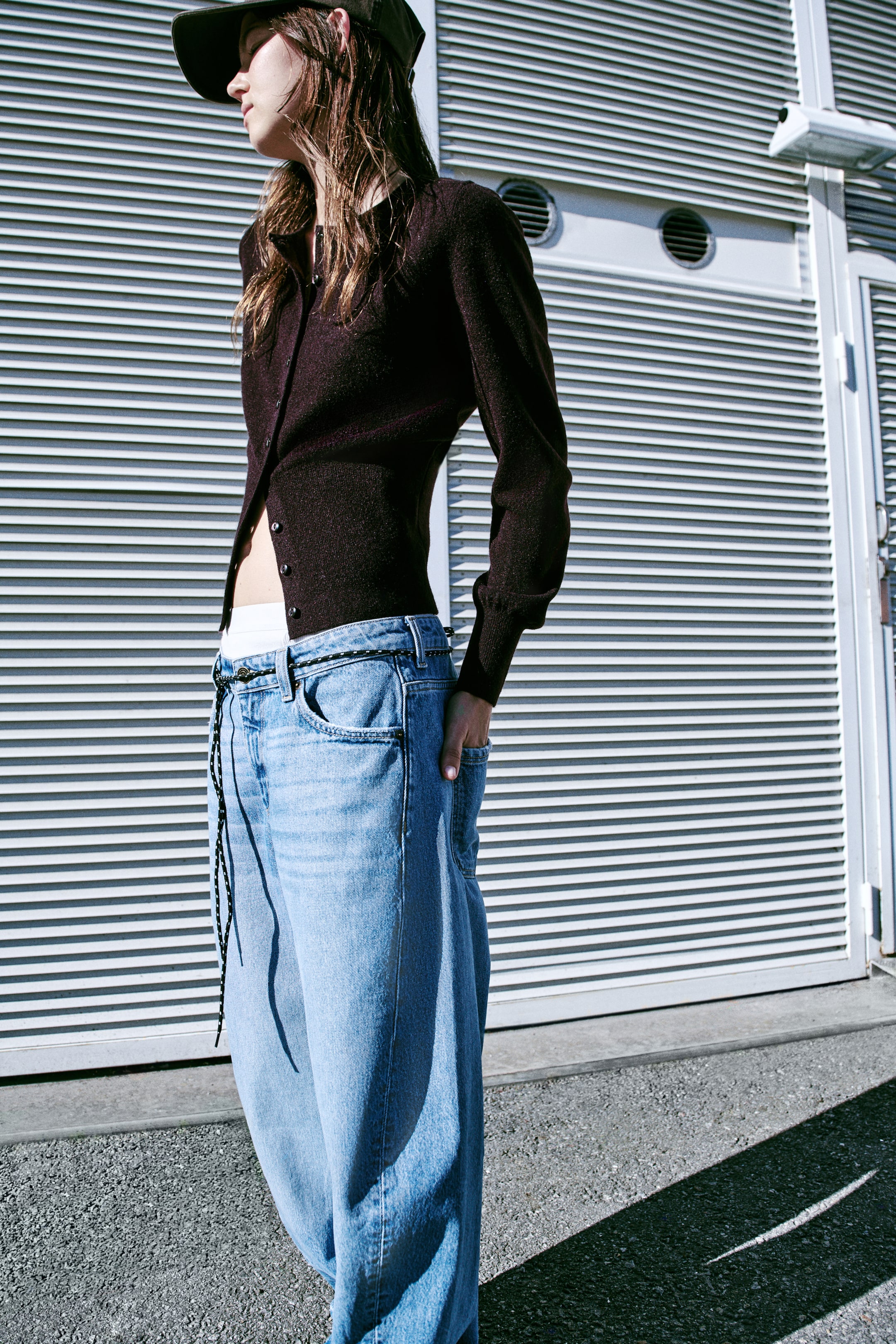 View larger image: A dark brown fine-knit cardigan is shown from a three-quarter front angle, featuring a round neckline, a button-down front, and long sleeves with wide ribbed cuffs. The cardigan is worn partially unbuttoned at the bottom, highlighting the wide ribbed hem and relaxed silhouette.