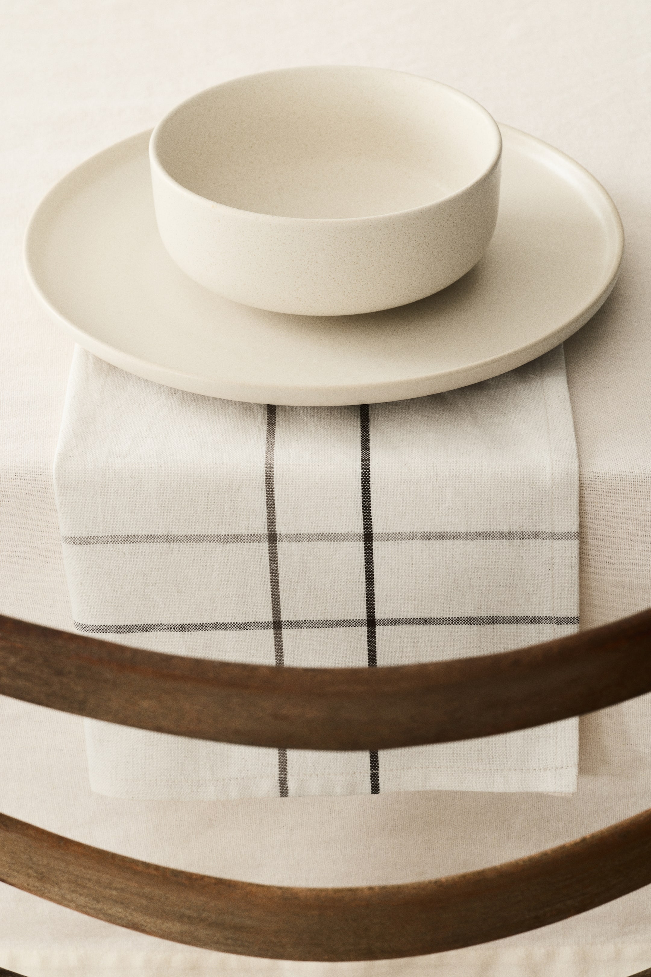 View larger image: A round, off-white, speckled dinner plate holds a matching round bowl, both pieces angled slightly upwards. The dinnerware rests on a folded, off-white napkin showcasing a dark grid pattern.