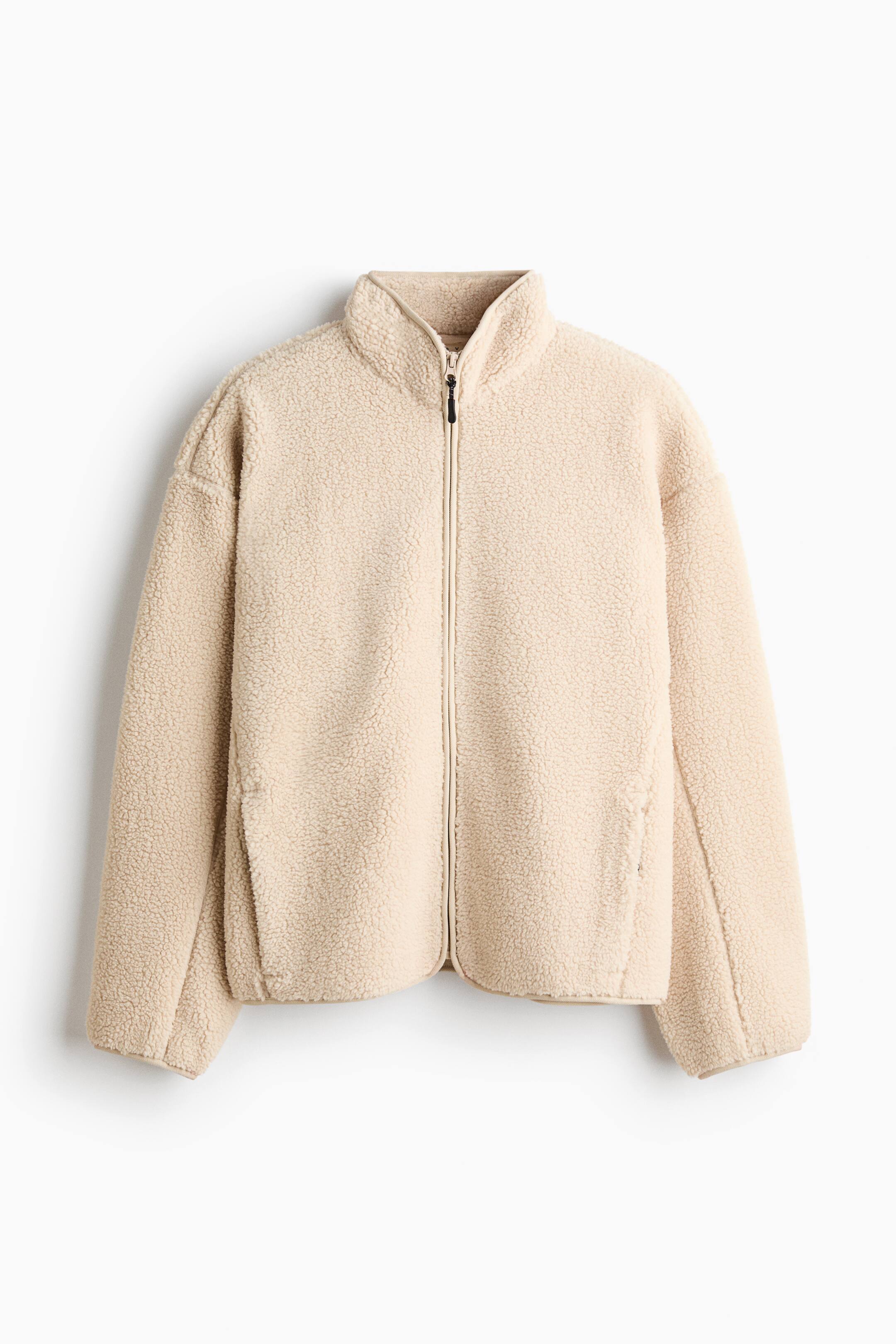 Se større billede: En beige sherpa-fleecejakke vises forfra med en tekstureret teddy-finish, høj opretstående krave og en gennemgående lynlåslukning. Jakken fremhæver en afslappet pasform med sænkede skuldre og diskrete sidelommer.