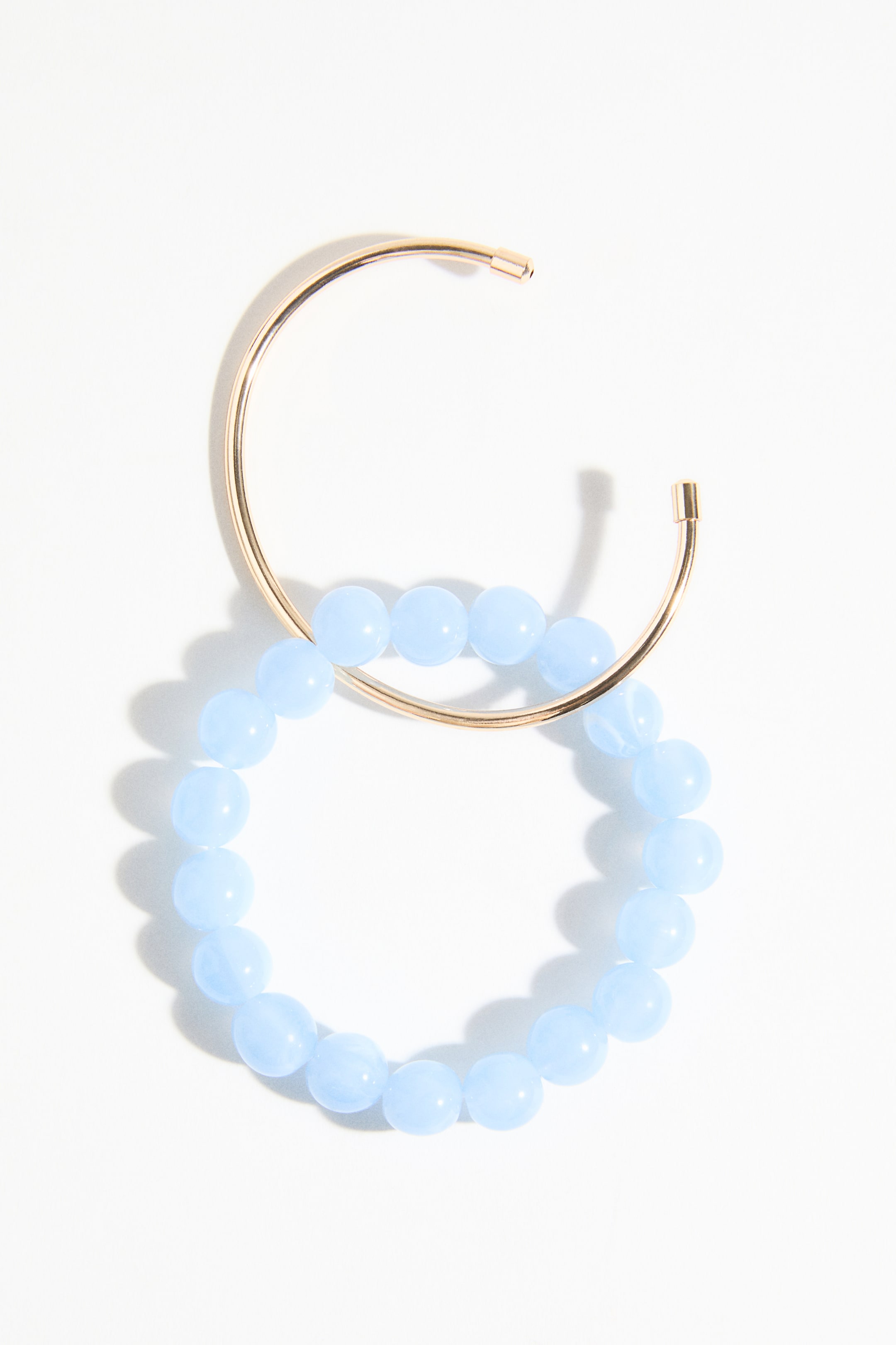 Ingrandisci l'immagine: Un bracciale a polsino aperto color oro sovrapposto a un bracciale con perline azzurro chiaro. Il polsino liscio a forma di U si curva intorno alle perline rotonde e opache azzurro chiaro, creando un cerchio completo.
