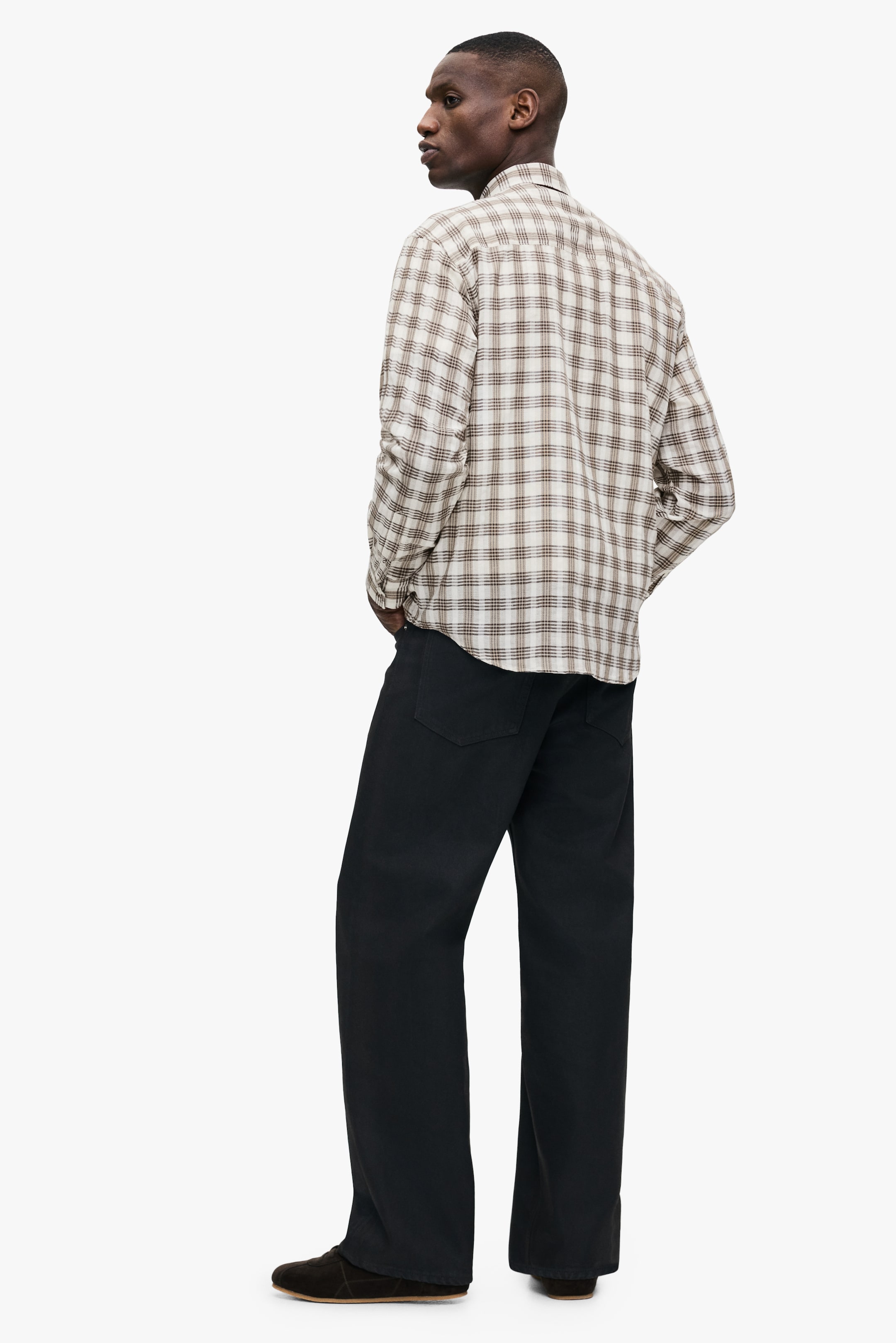 查看大圖: 模特穿著一件長袖格子襯衫，襯衫以乳白色為底，配有棕色和黑色的格子，沒有紮進褲子裡，垂落在黑色長褲上。