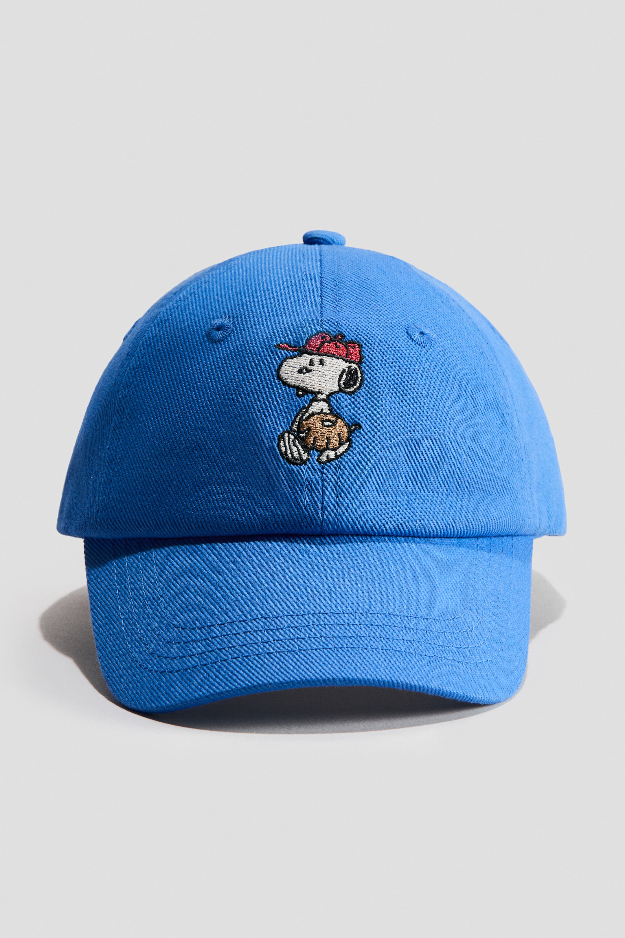 View larger image: A blue cotton twill cap showcasing an embroidered motif of Snoopy wearing a red cap and holding a brown baseball glove on its front. Embroidered eyelets are visible on the crown.