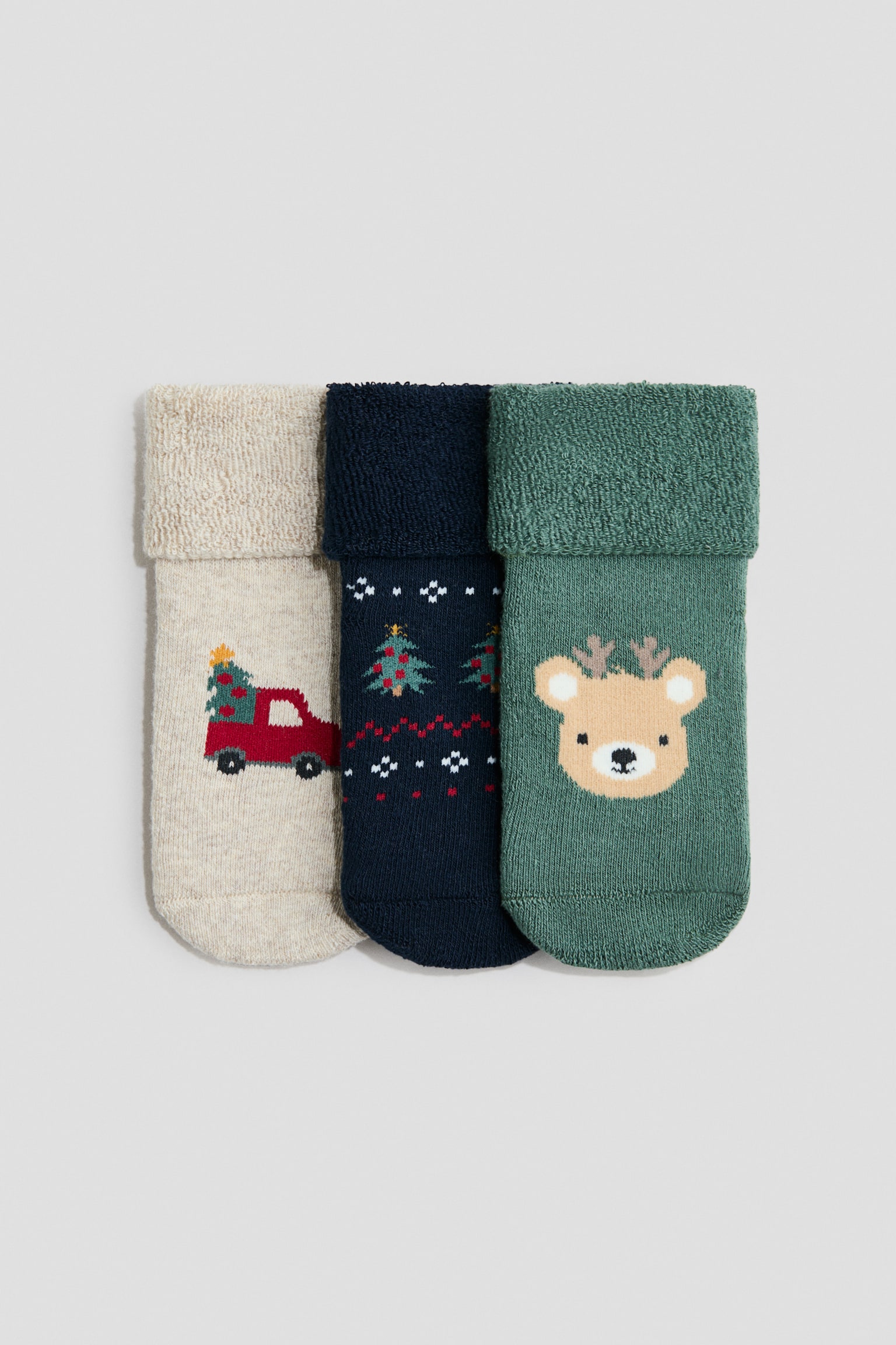 Tre finstrikkede sokker ligger fladt og vender for, og viser opsmÃ¸gede skaft med en struktureret frottÃ©foring. Sokkerne fremviser tydelige intarsia-designs med juletema, herunder en rÃ¸d lastbil med et trÃ¦ pÃ¥ den beige sok, festlige mÃ¸nstre pÃ¥ den marineblÃ¥ sok og en bjÃ¸rn med gevir pÃ¥ den grÃ¸nne sok.