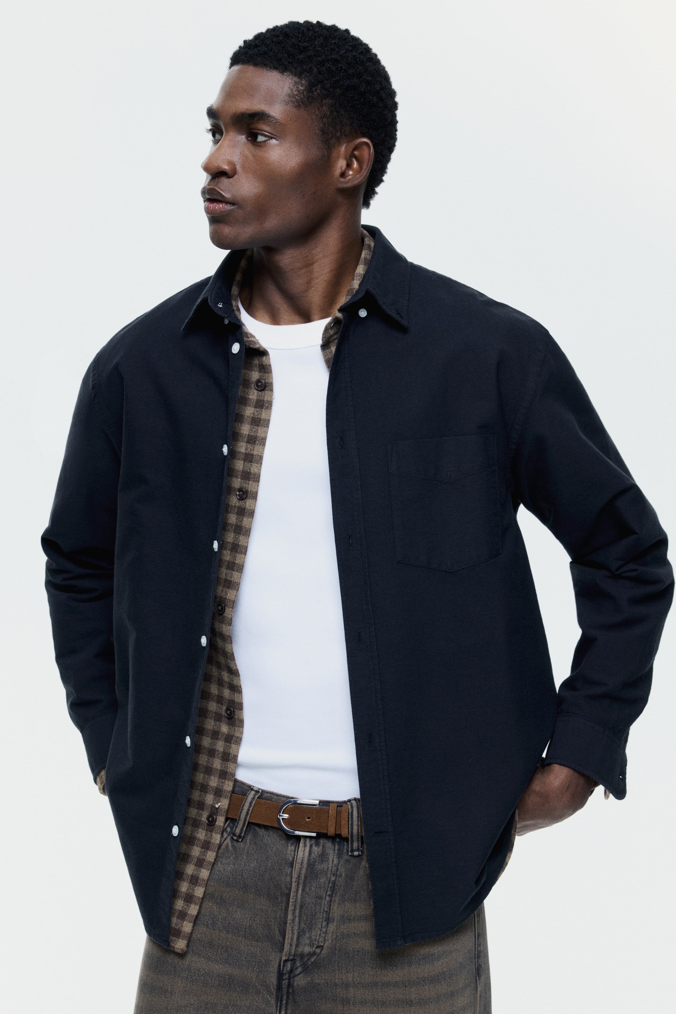View larger image: The image showcases a black collared shirt from a slight front-right perspective, highlighting its long sleeves and a single left chest pocket. The shirt is styled open to reveal a brown and white plaid button-up shirt beneath, layered over a white ribbed t-shirt.