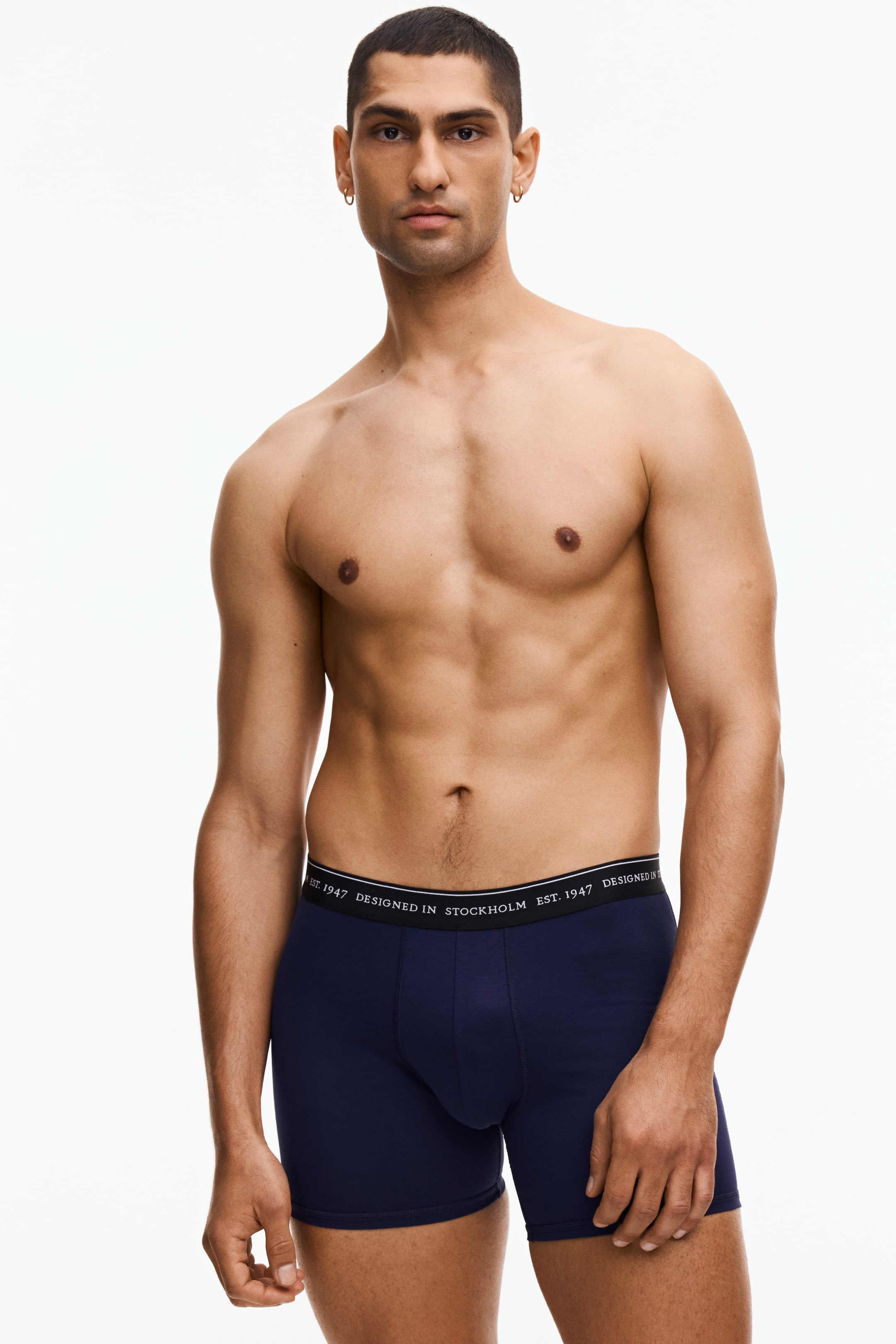 View larger image: Navy blue boxer briefs are being worn, featuring a black elastic waistband with repeating white text 'EST. 1947 DESIGNED IN STOCKHOLM' and a contoured front pouch, shown from a slightly front-right angle.