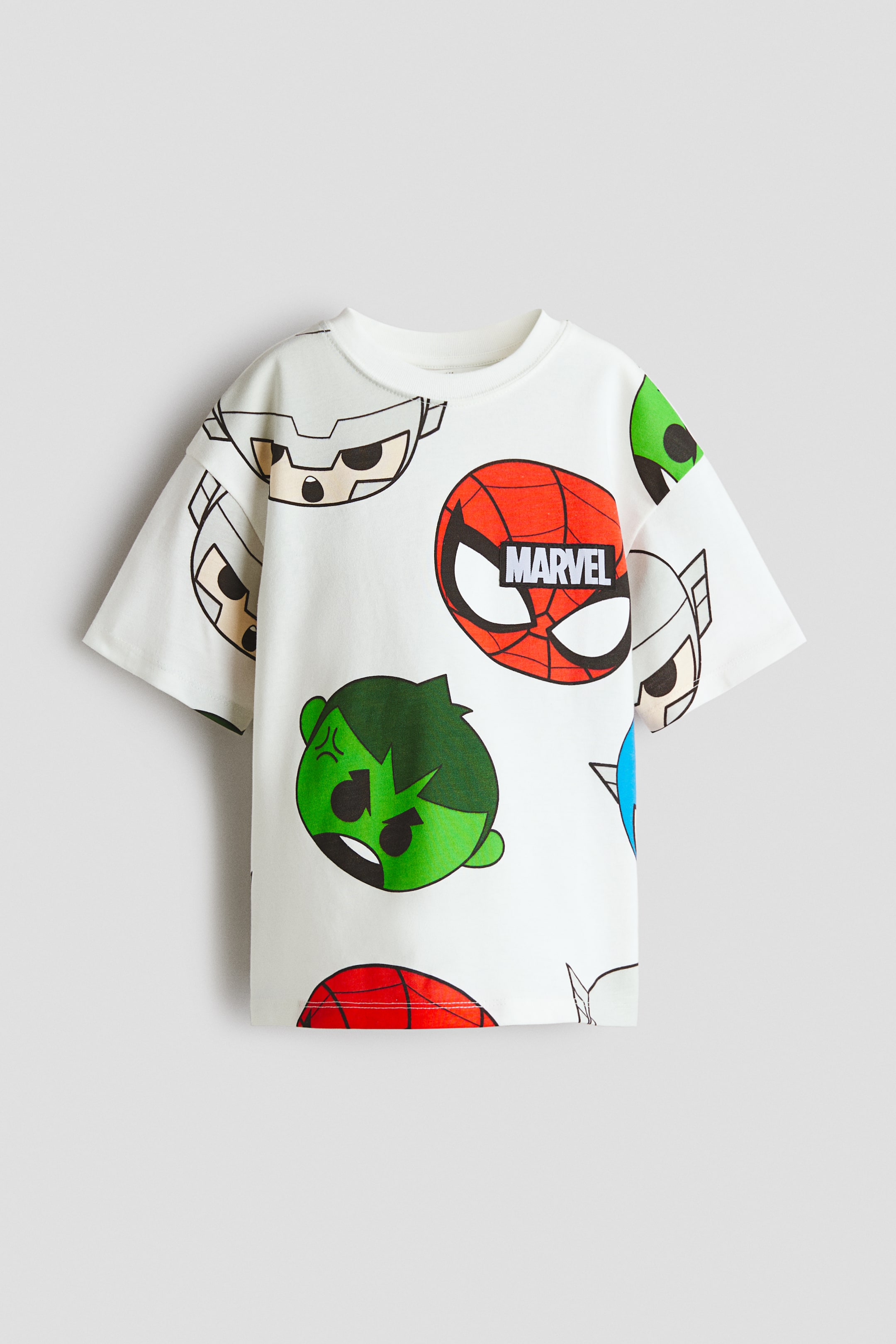 Agrandir l'image: Un t-shirt à coupe ample est présenté de face, avec un imprimé all-over de visages de personnages Marvel stylisés et colorés sur fond blanc cassé. Il est conçu avec un col rond côtelé et des épaules tombantes.
